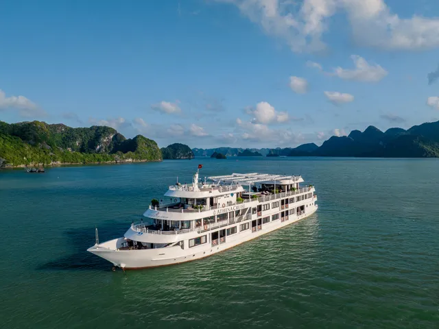 Aerial view of a white multi-deck cruise ship with 'Athena Cruise' lettering on its side and a Vietnamese flag, sailing on green water among forested limestone karst islands under a blue sky.