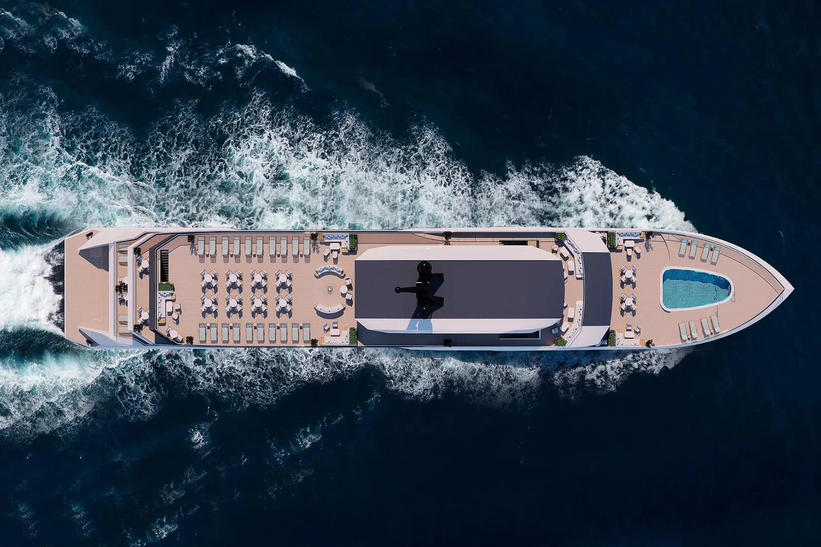 A top-down aerial view of a large cruise vessel with a sun deck, lounge chairs, tables and a small pool at the bow, moving through dark blue open water with a white wake behind it.