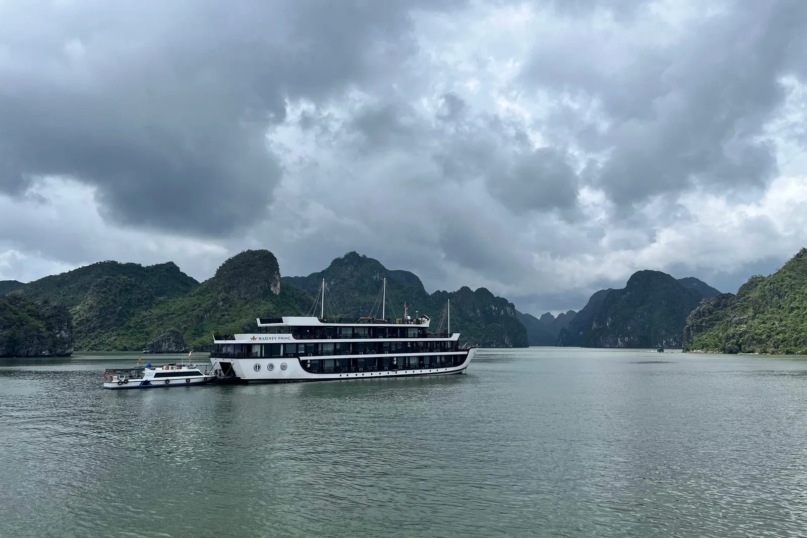 A multi-deck white cruise ship with 'Majesty Prime' lettering on its hull sits on calm green water beside a small tender boat, with forested limestone karst islands behind and a heavy overcast sky above.