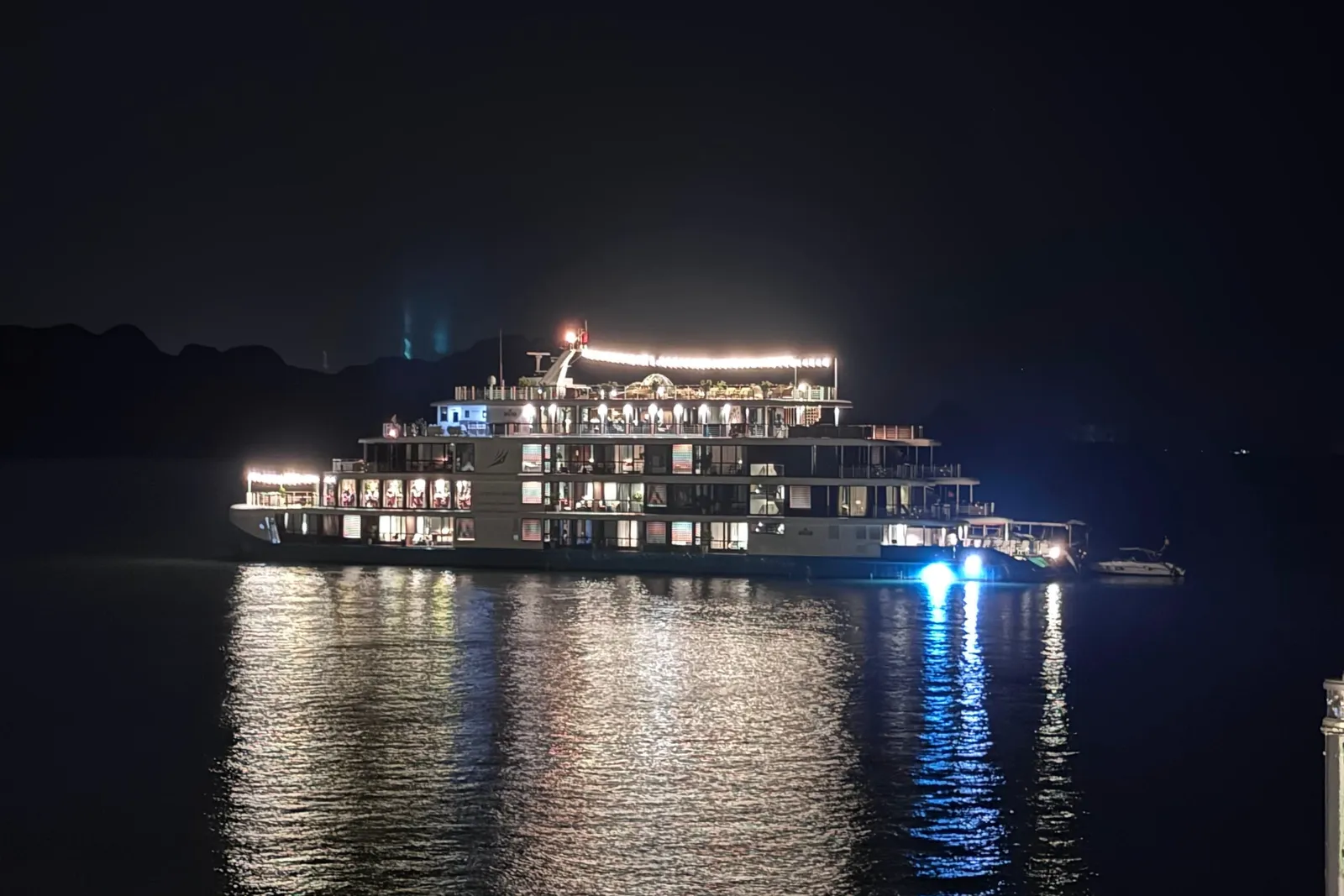 A multi-deck cruise ship lit up at night on dark water, with a small boat beside it, dark hills in the background, and a railing post at the right edge of the frame.