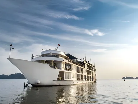 A large white multi-deck cruise ship with gold anchor emblems on its balconies sits on calm water, seen from the bow side, with distant limestone islands and a blue sky with wispy clouds behind it.