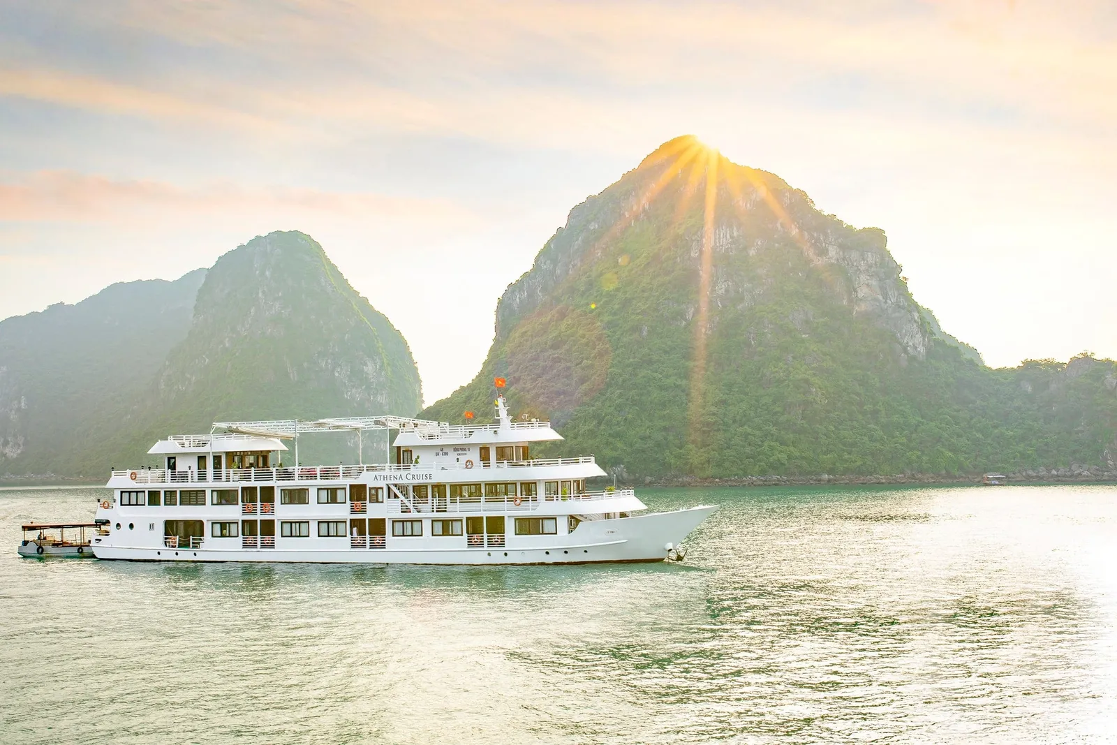 A white multi-deck cruise boat with 'ATHENA CRUISE' lettered on its side sails on calm green water in front of forested limestone karst peaks with the sun bursting over the summit.