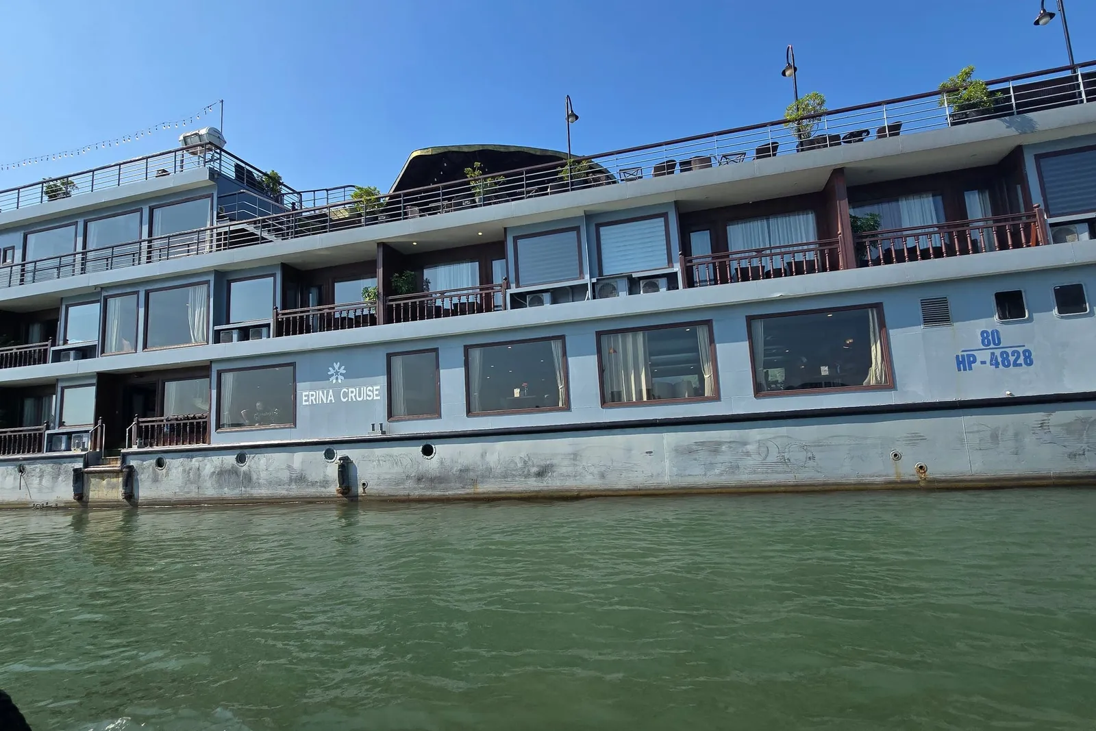 A multi-deck grey cruise ship with the painted name 'ERINA CRUISE' and registration 'HP-4828' on its hull, photographed from a small boat at water level on green water under a clear blue sky.