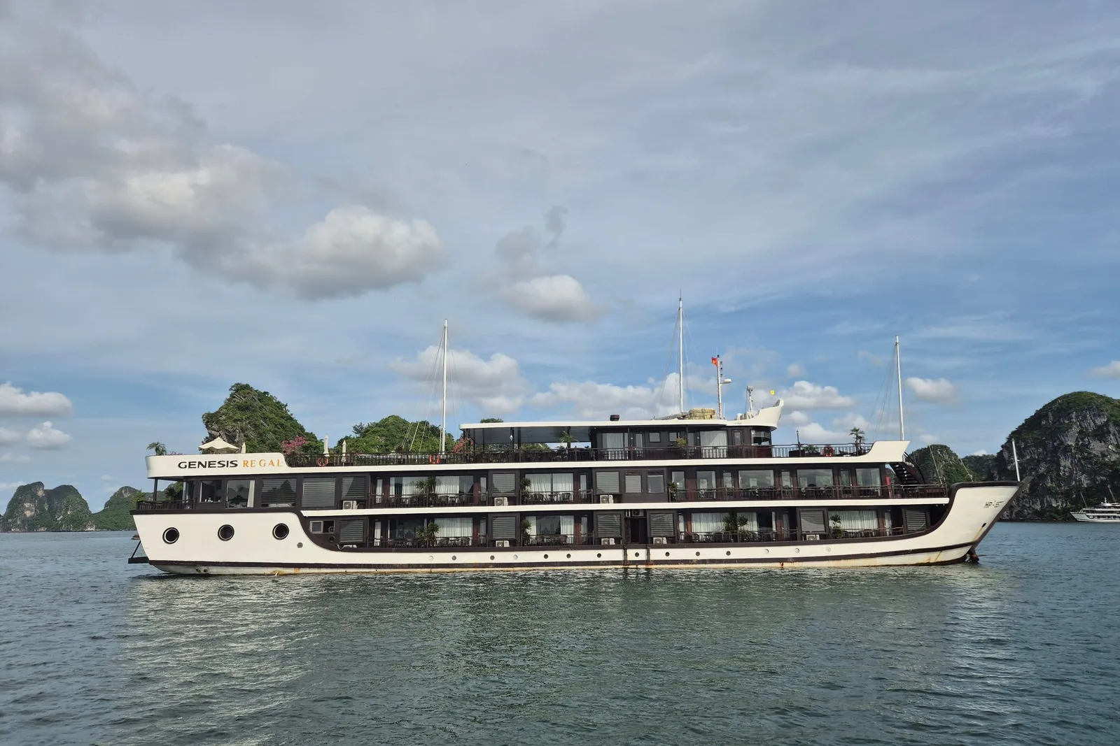 A white and dark-brown multi-deck cruise ship with 'Genesis Regal' lettering on its upper hull sits on calm green water in front of limestone karst islands under a partly cloudy sky.