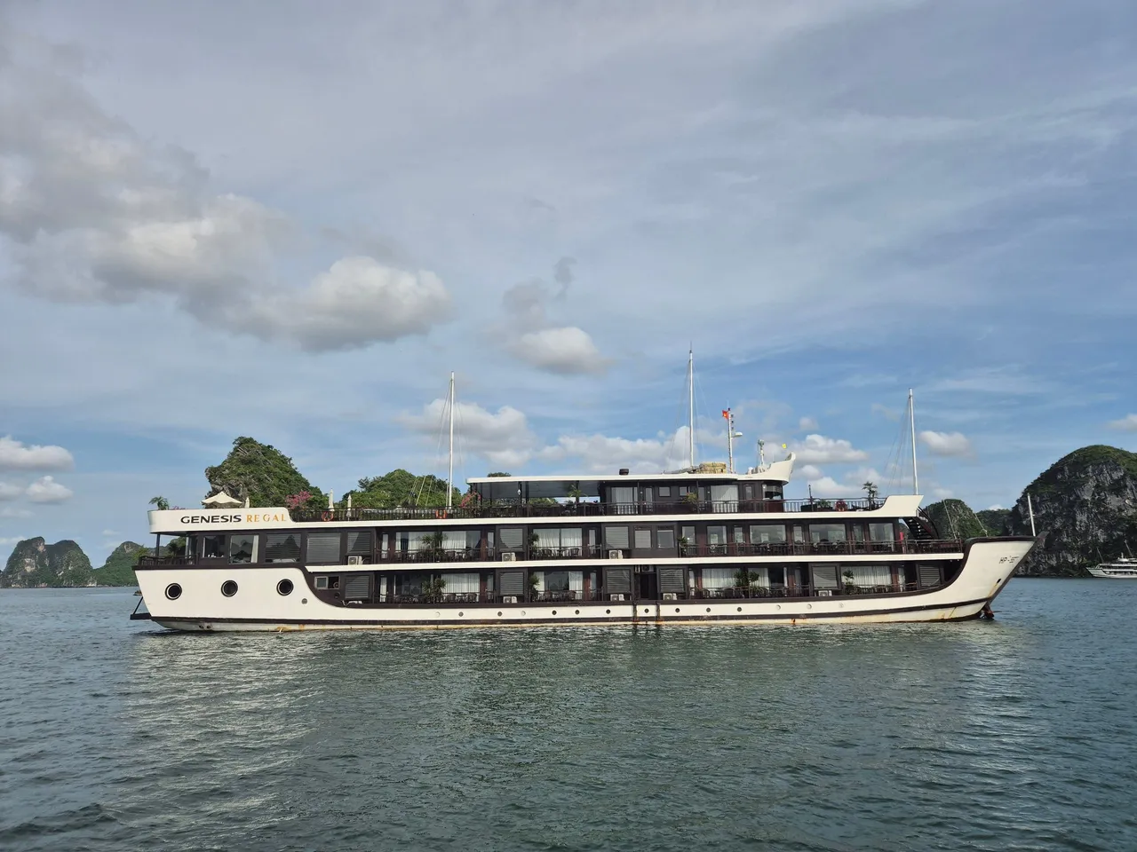 A white and dark-brown multi-deck cruise ship with 'Genesis Regal' lettering on its upper hull sits on calm green water in front of limestone karst islands under a partly cloudy sky.