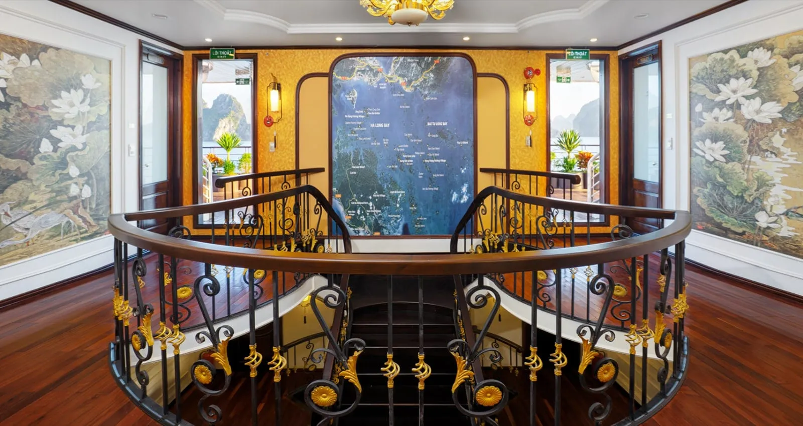 Interior of a stairwell landing with a curved wooden handrail and ornate black and gold wrought-iron balustrade, a framed Ha Long Bay map on the back wall, lotus paintings on both side walls, and glass doors opening to a balcony with karst views.
