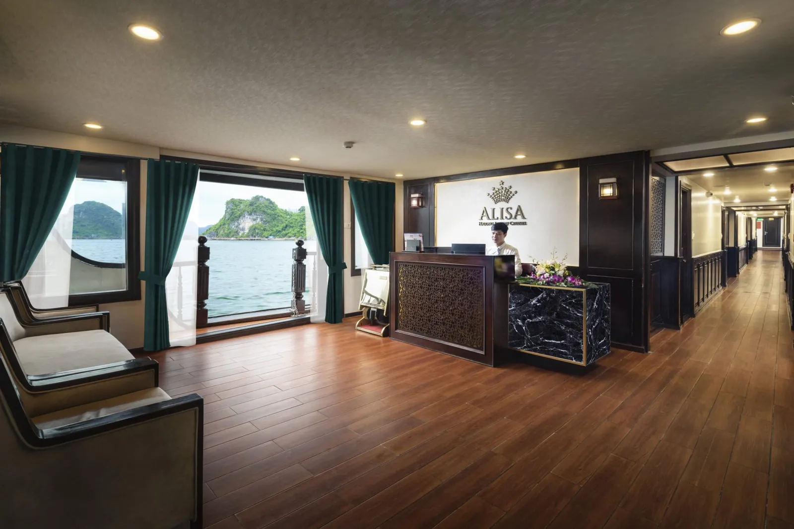 Interior reception lobby of a cruise boat with a staff member at a wooden desk beneath a wall logo reading 'ALISA', lounge chairs on the left, a long wood-floored corridor on the right, and windows showing green karst islands on the water.