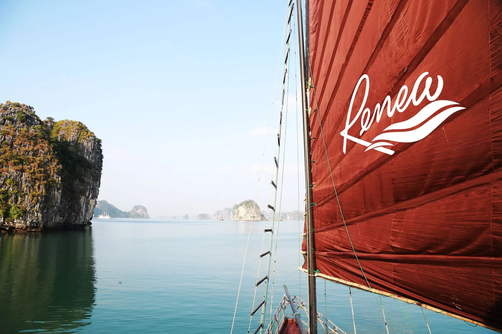 View from the bow of a junk boat past a red sail printed with the white 'Renea' logo, across calm green water toward limestone karst islands under a hazy blue sky.