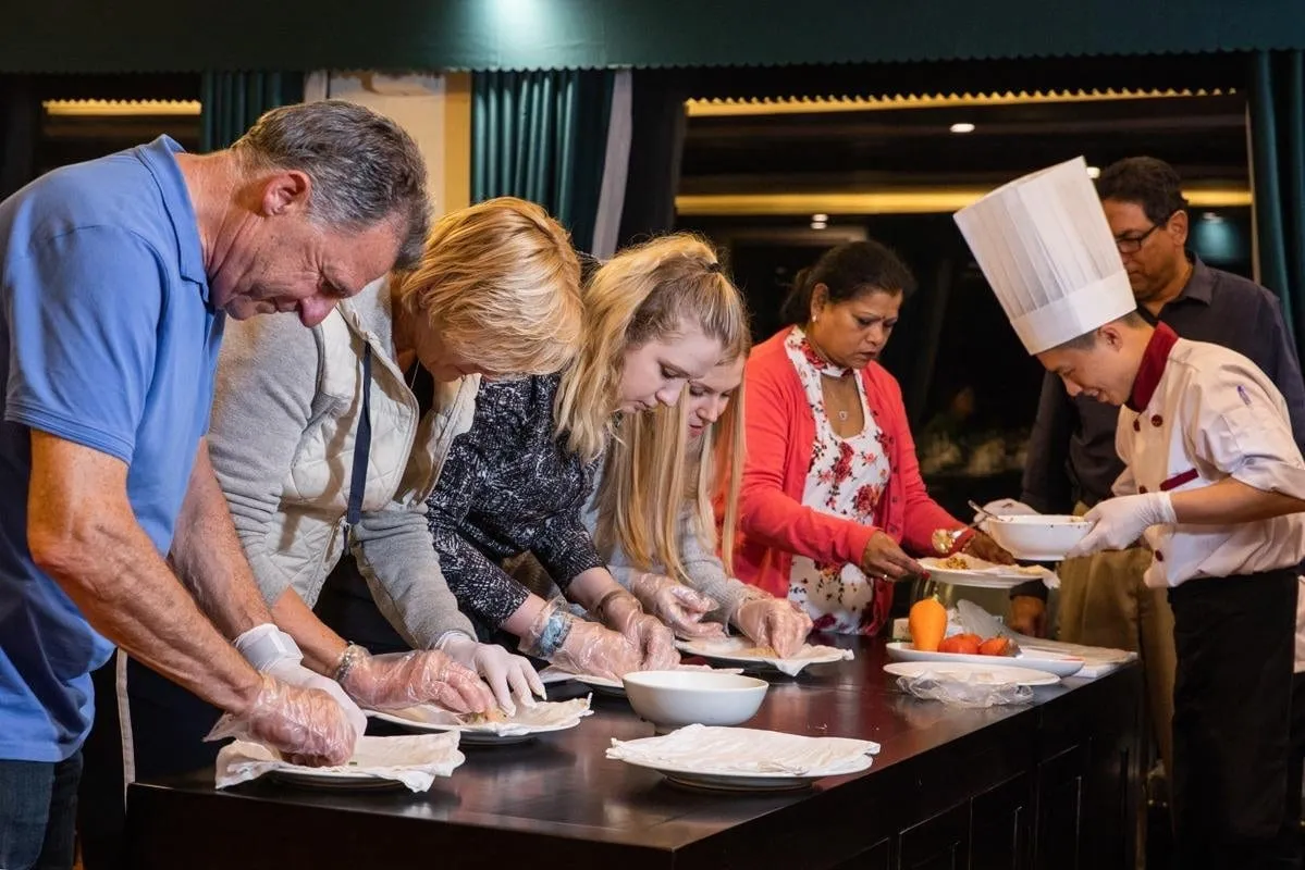 Guests wearing clear plastic gloves prepare food at a dark counter while a chef in a tall white hat assists, in a dim indoor room with green curtains.