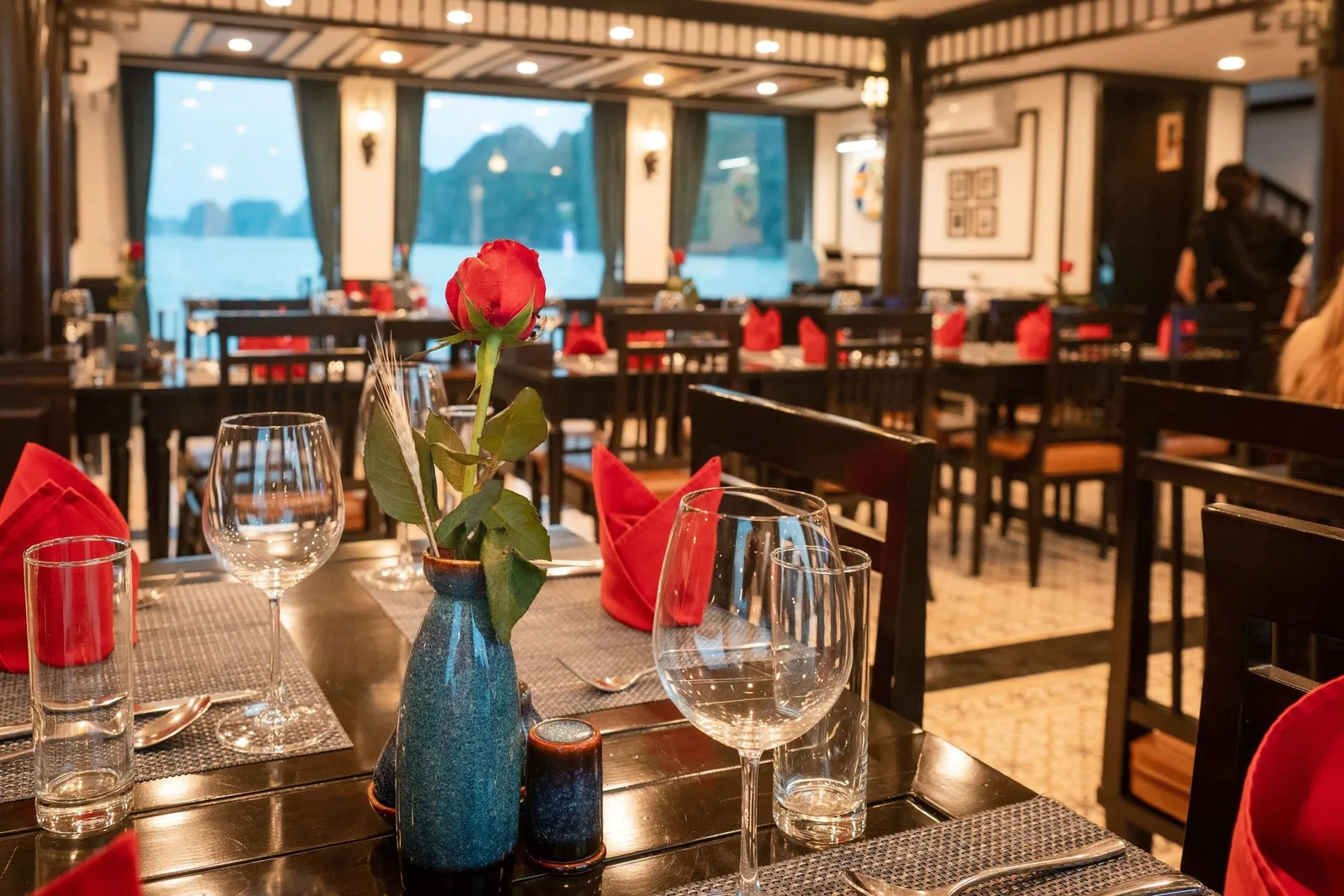A dining room with set tables, red napkins, wine glasses and a red rose in a blue vase, with large windows showing misty limestone karst islands on the water.