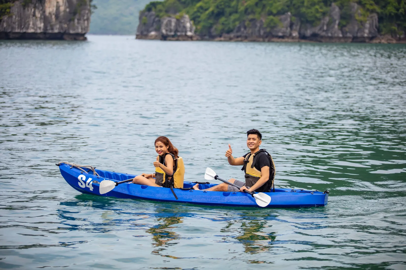 A woman and a man in yellow life vests paddle a blue tandem kayak marked "S4" on calm green water, with the man giving a thumbs up and limestone karst islands in the background.