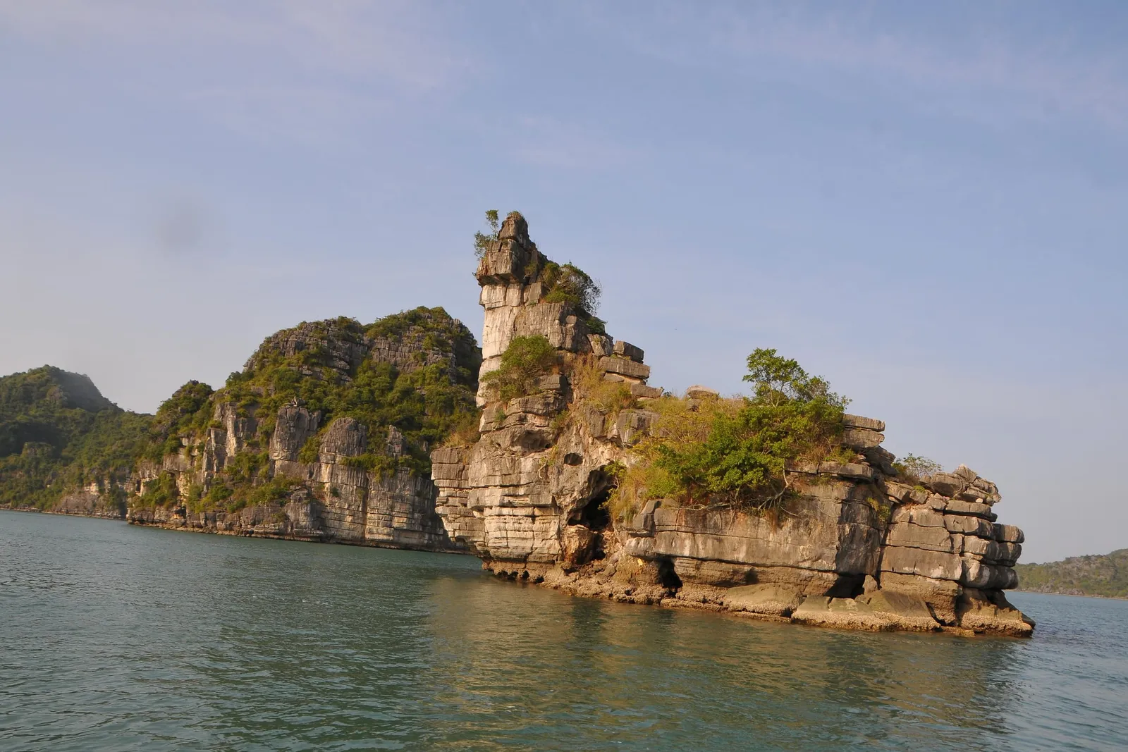 A limestone rock formation with a tall pillar and green vegetation rising from calm green water, with forested karst hills behind and a pale blue sky, taken from water level.