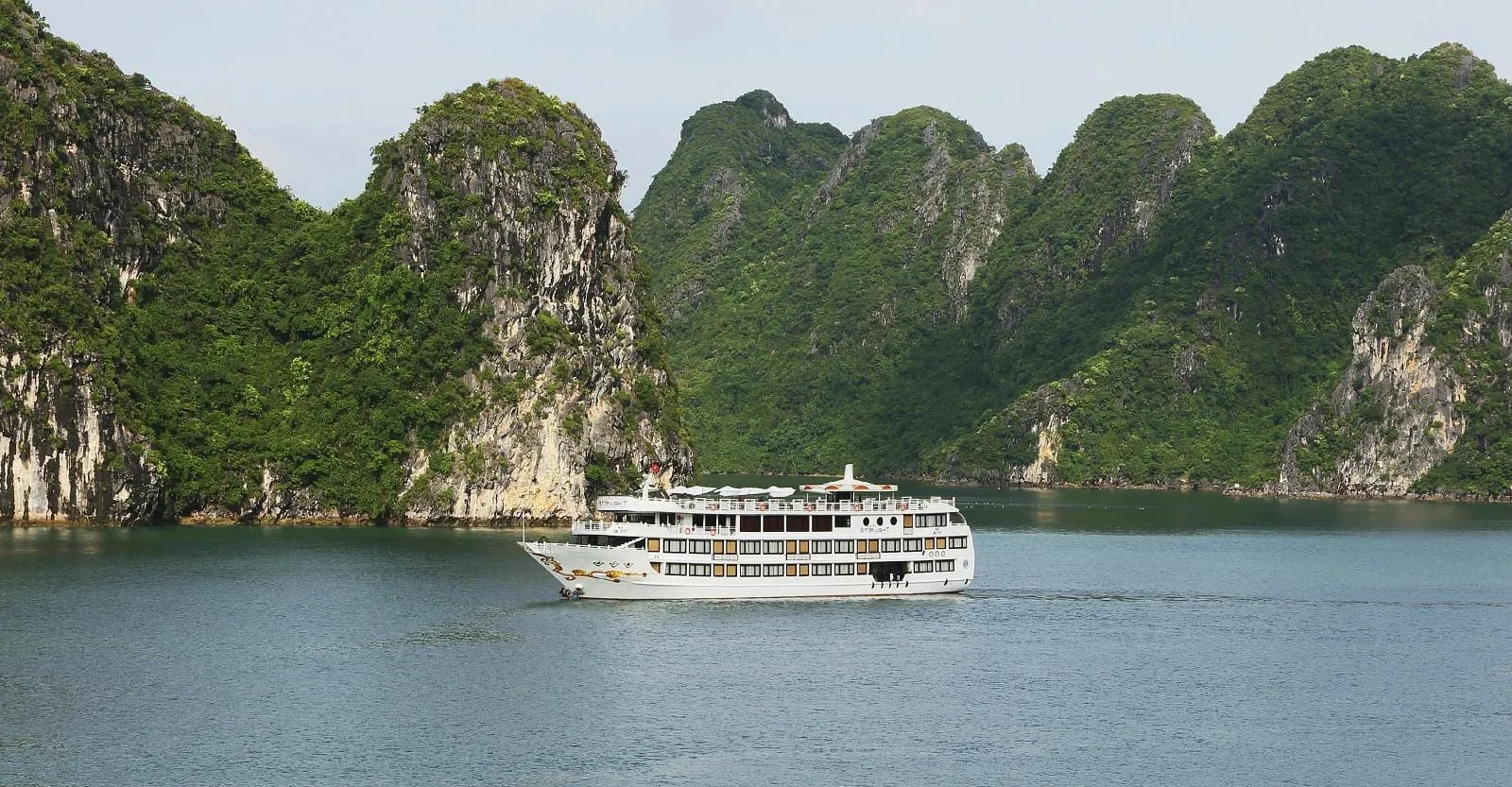 A white multi-deck cruise boat sits on calm green-grey water in front of steep, forest-covered limestone karst islands under a pale sky.