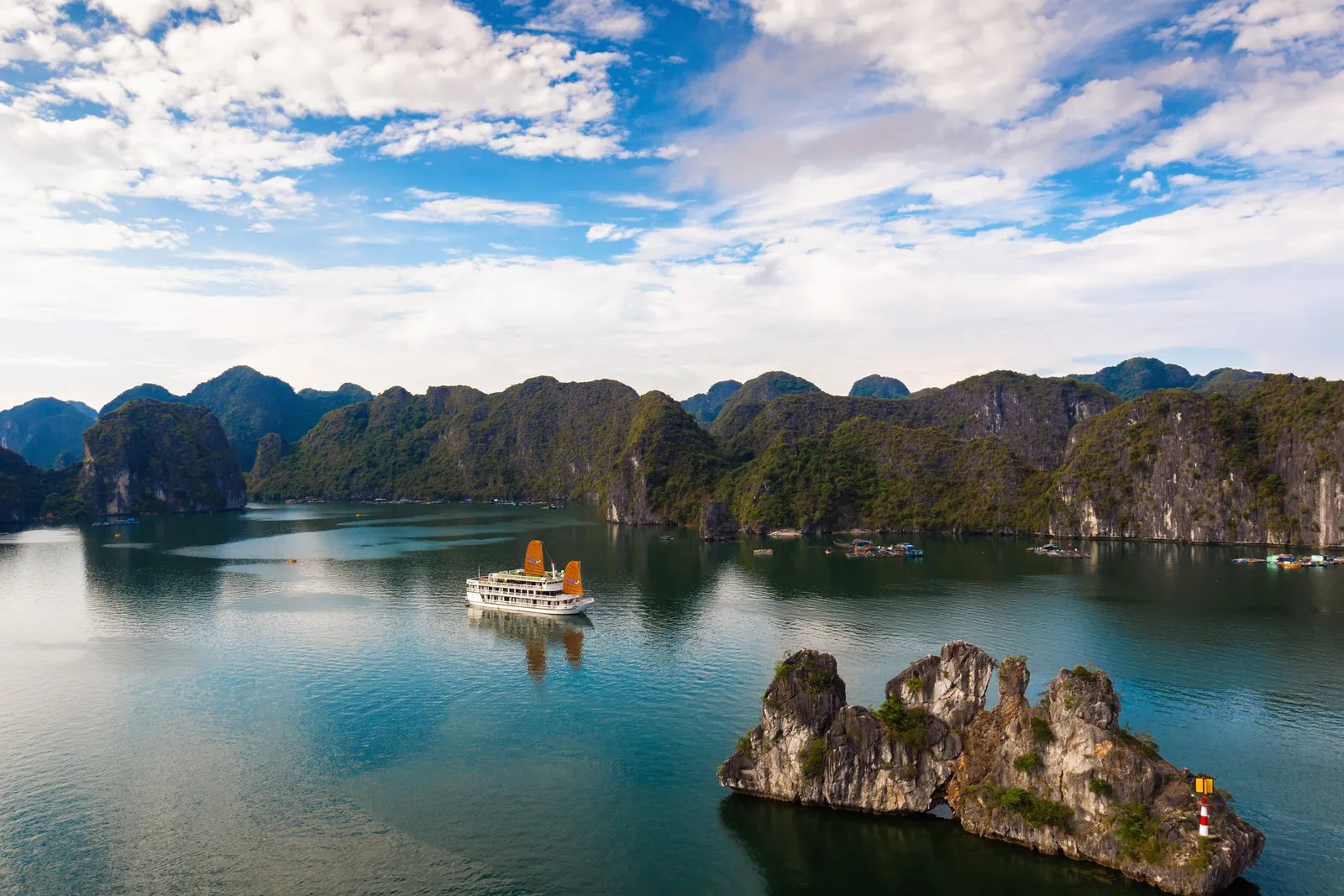 An aerial view of a white cruise ship with orange sails on calm teal water, with a rocky islet topped by a small red-and-white lighthouse in the foreground and green karst mountains under a blue, cloudy sky.