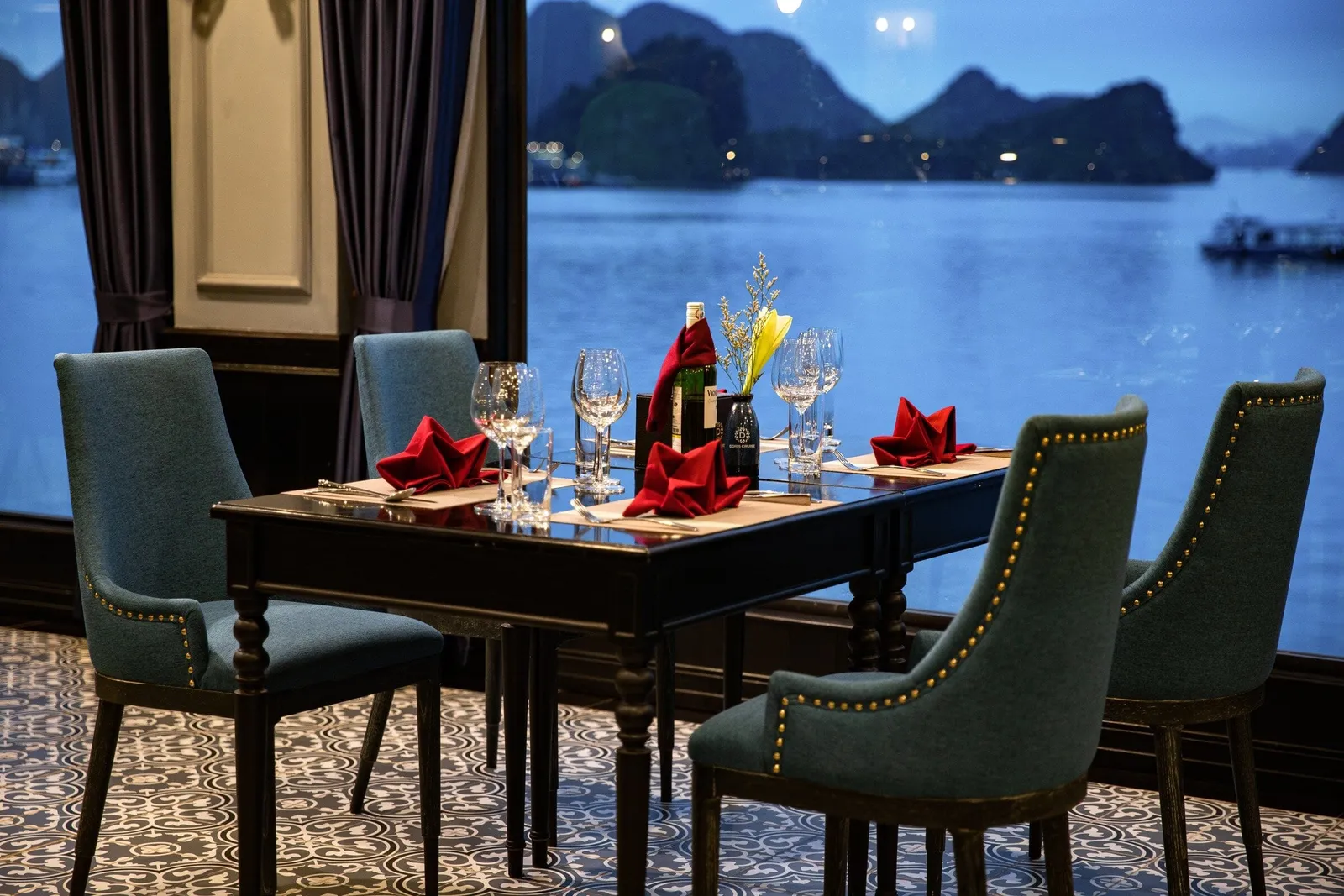 A dining table set with red folded napkins, wine glasses and a flower vase, surrounded by teal studded chairs, beside a large window overlooking a blue bay with limestone karst islands at dusk.