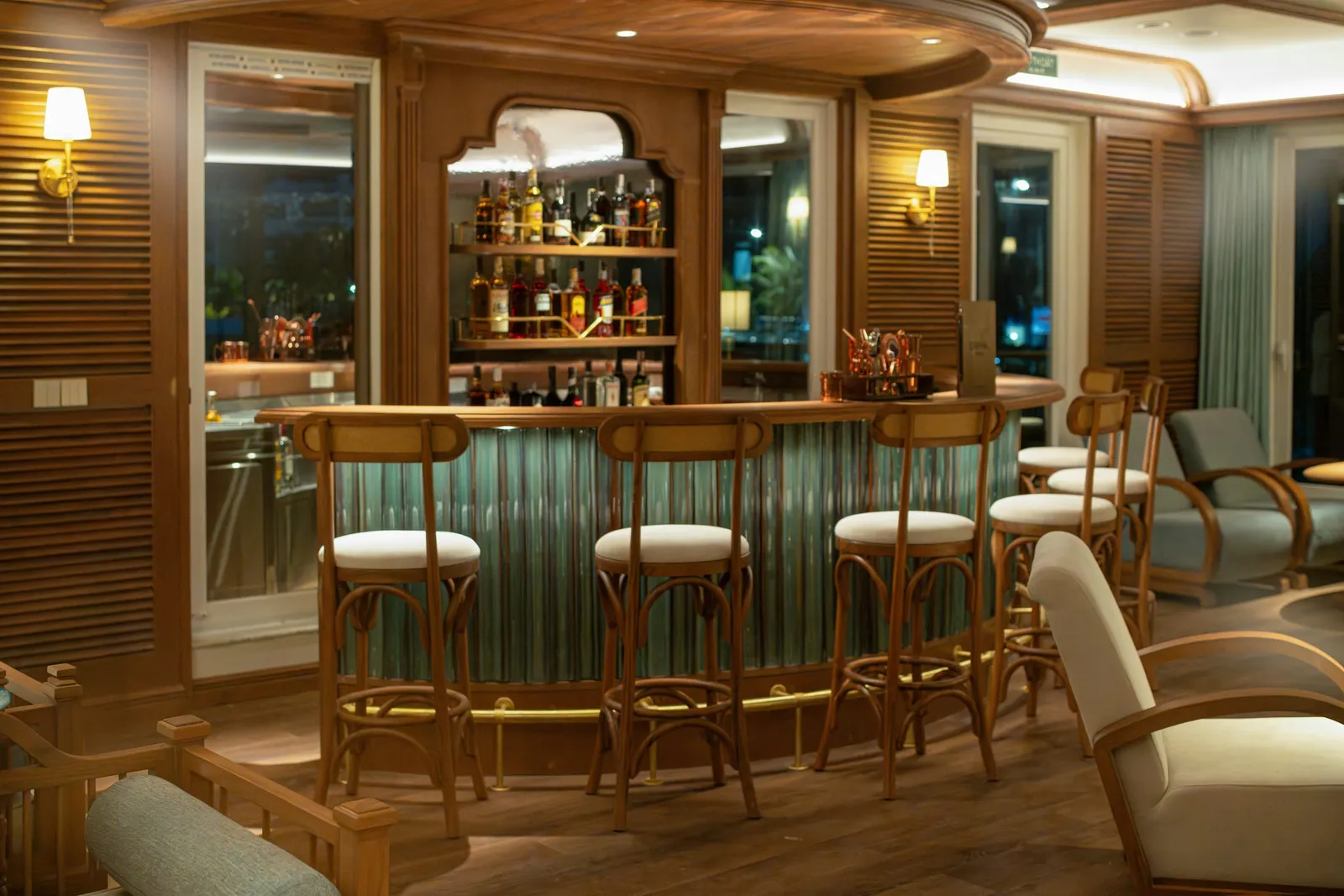 A curved wooden bar with teal fluted front, tall stools, shelves of liquor bottles, wall lamps, louvered wood panels and armchairs in a warmly lit lounge, with night-time lights visible through the windows.