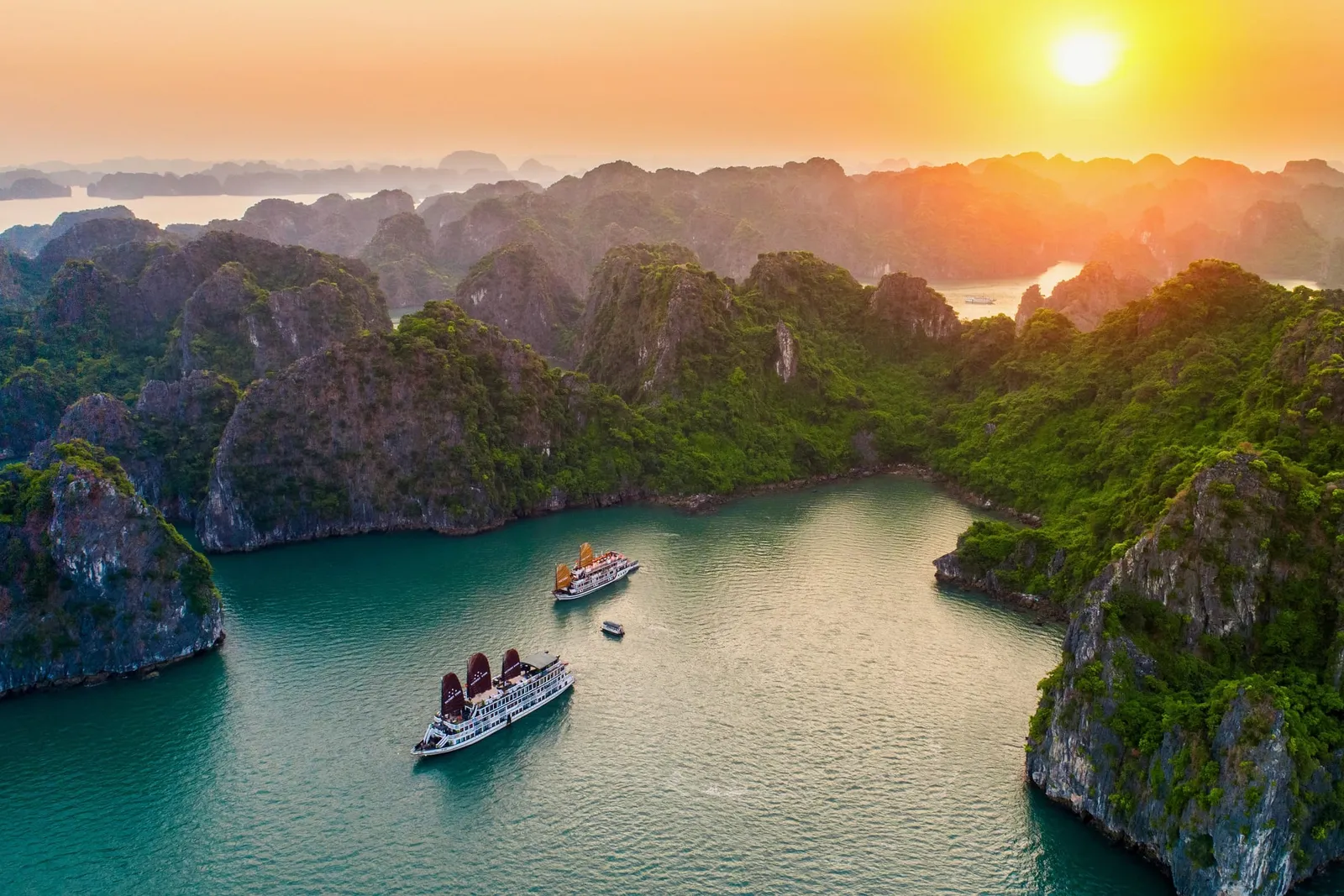 Aerial view at sunset of two white cruise boats, one with dark red sails, on turquoise water among green limestone karst islands.