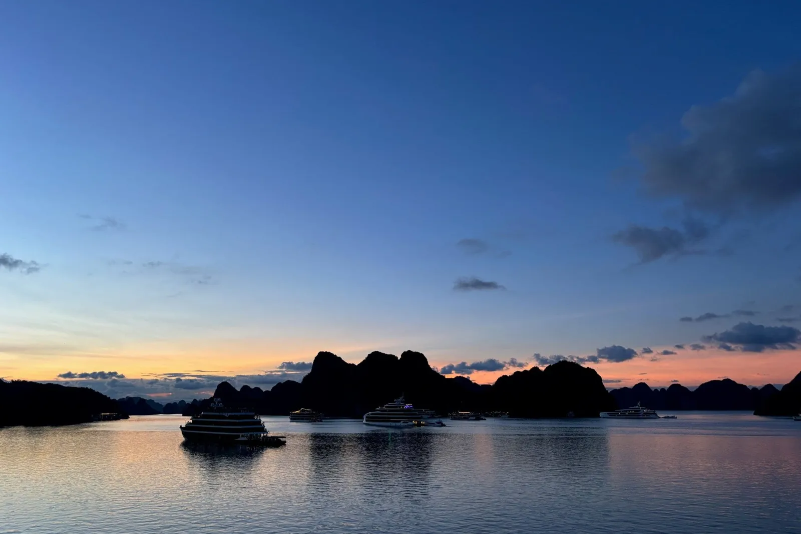 Dusk view across calm water toward silhouetted limestone karst islands and several anchored cruise boats under a blue-to-orange sky, with a railing edge visible at the bottom left.