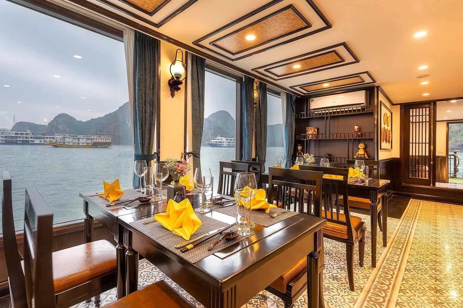 Interior of a cruise boat dining room with dark wooden tables set with yellow folded napkins and glasses, beside large windows overlooking calm water, other boats, and limestone karst peaks.