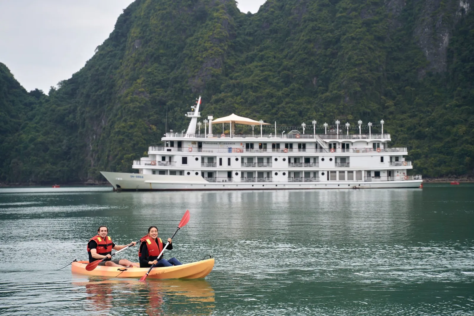 Two people in orange life vests paddle a yellow kayak on green water in front of a large white multi-deck cruise ship, with forested limestone karst hills behind.