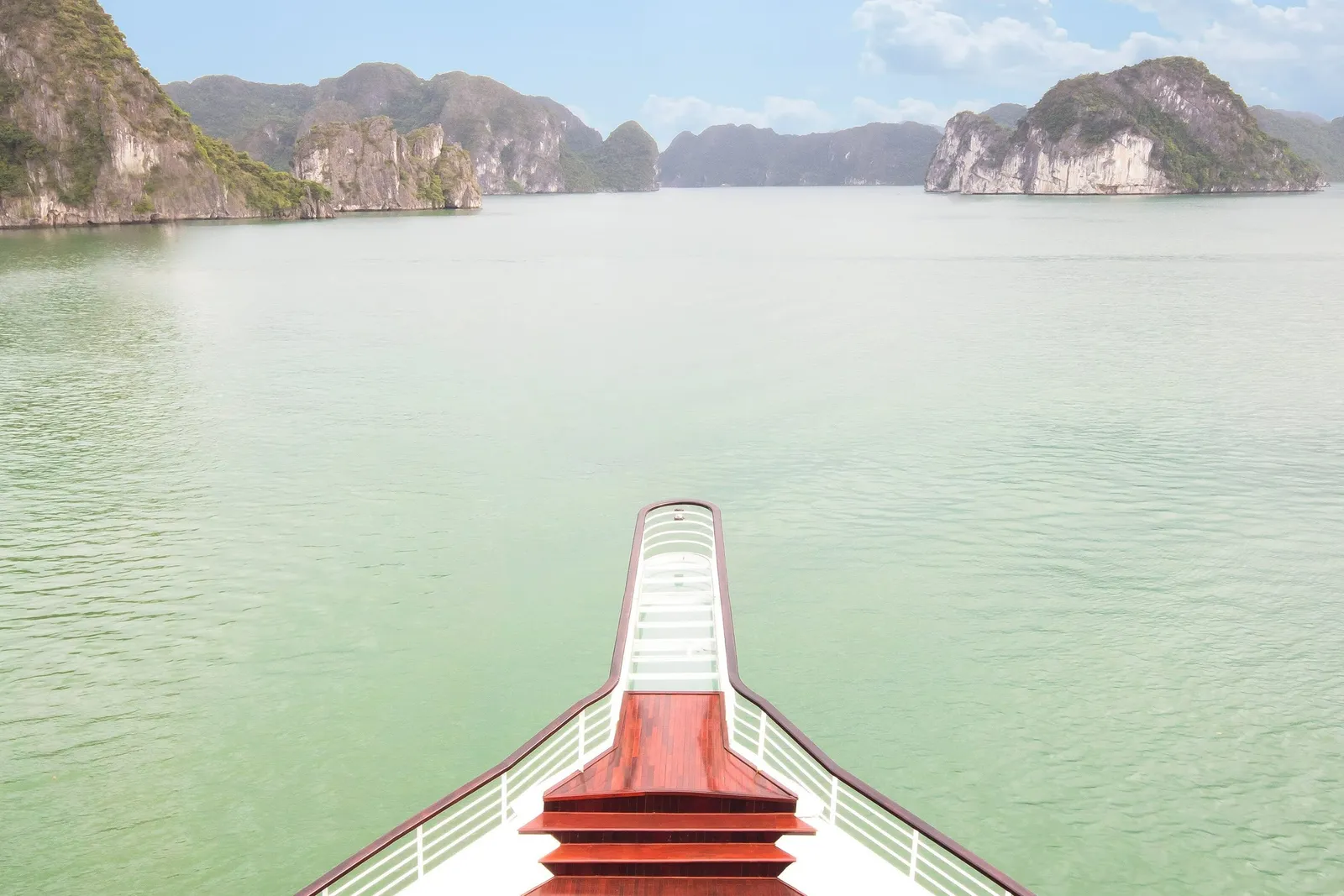 View from an upper deck over the bow of a boat with wooden steps and white railings, looking out across calm green water toward limestone karst islands under a hazy blue sky.