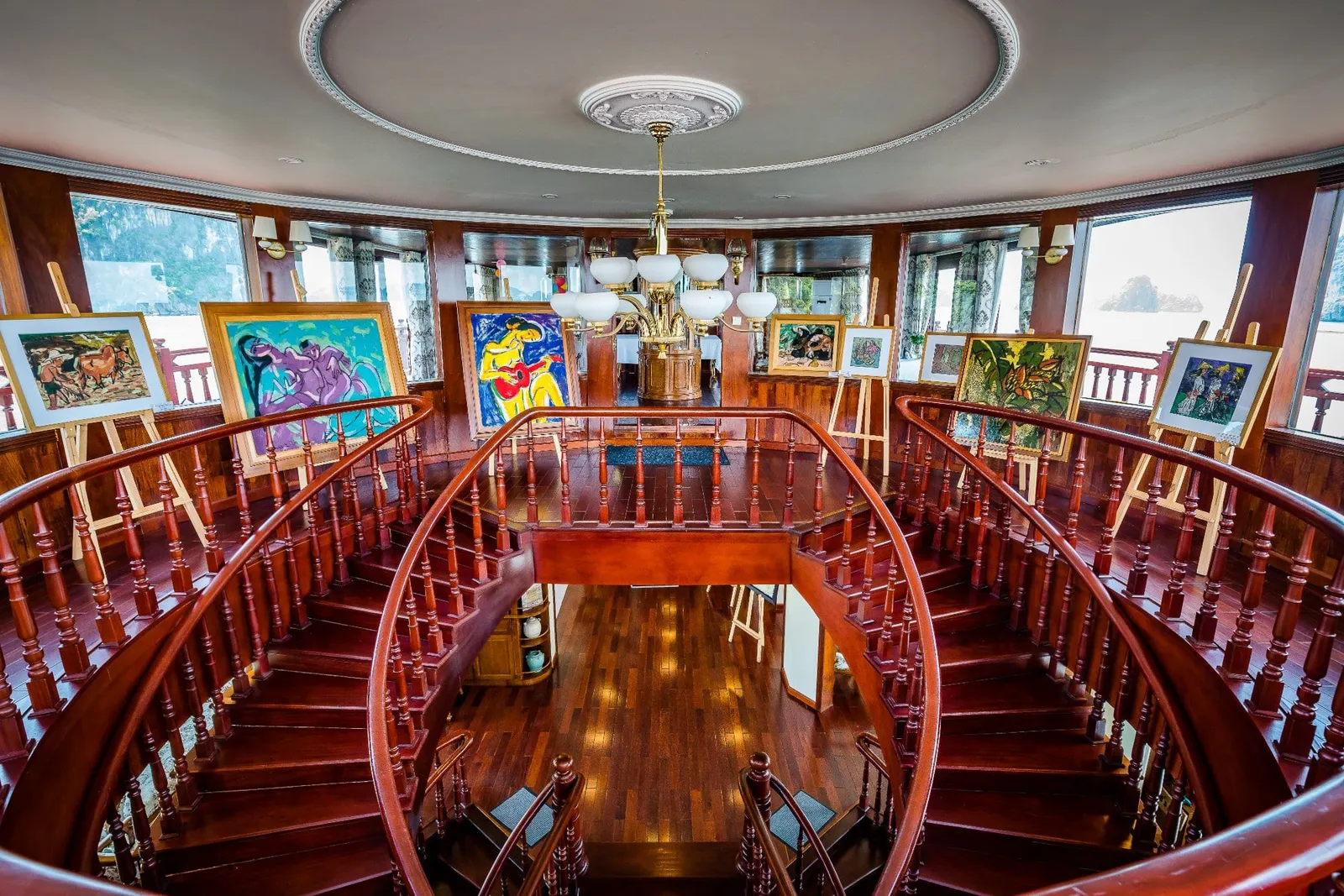 A curved double wooden staircase with a chandelier and paintings on easels in a round, wood-paneled interior, with windows showing misty water and a karst island outside.