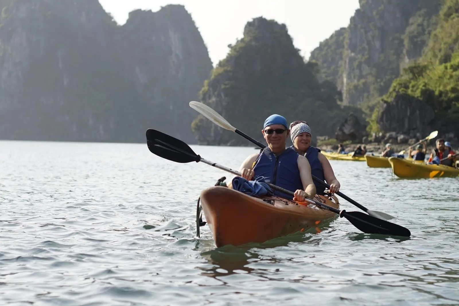Two people in life vests paddle a tandem kayak on calm green water with more kayaks behind them and forested limestone karst cliffs in the background.