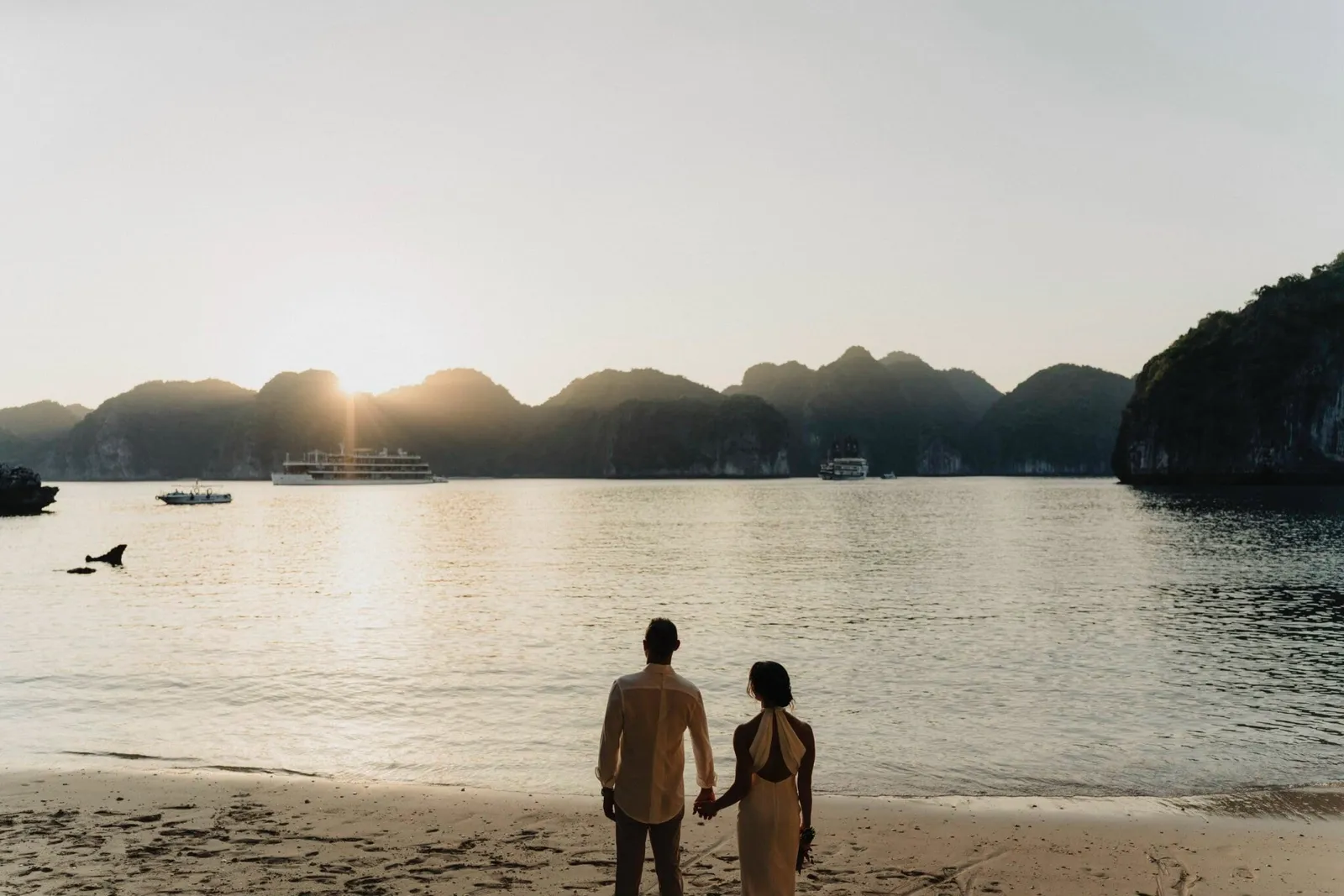 A couple holding hands, seen from behind on a sandy beach, looking out at a calm bay with limestone karst islands, cruise boats, and the sun setting behind the hills.