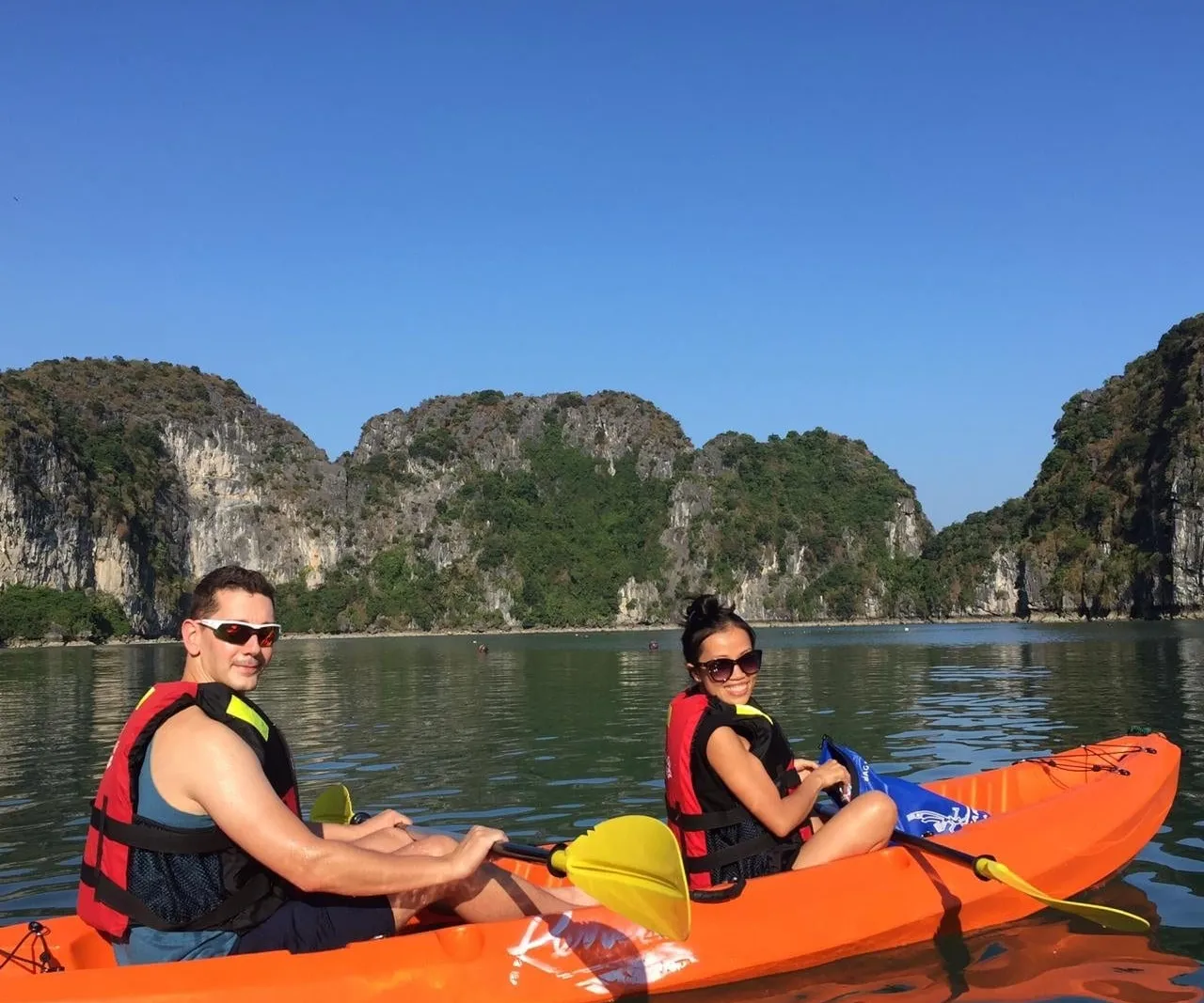A man and a woman in red life vests sit in an orange tandem kayak with yellow paddles on calm green water, with limestone karst islands and a clear blue sky behind them.