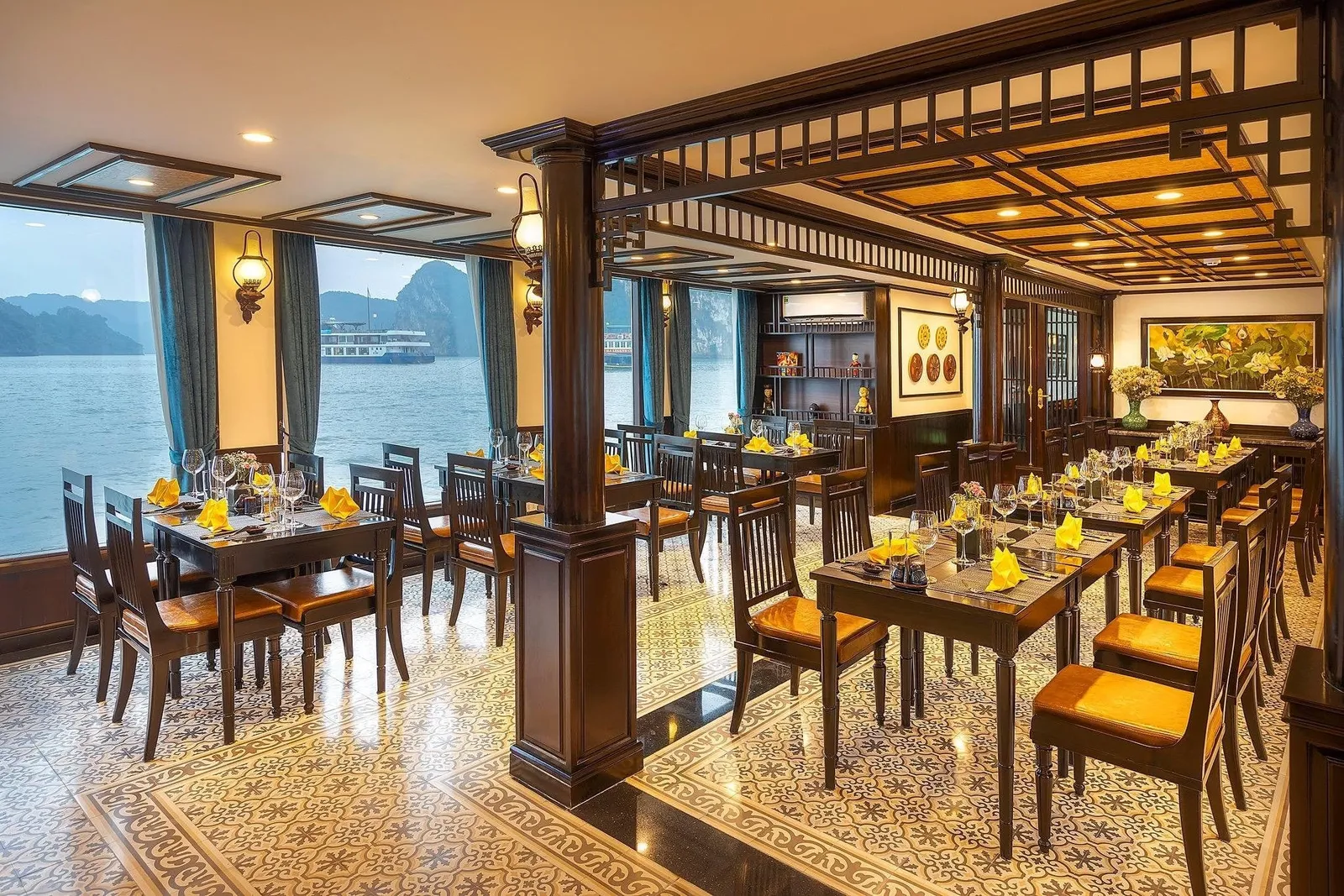 Interior of a cruise ship dining room with set wooden tables, yellow napkins and patterned tile floor, with windows showing limestone karsts and another boat on the water.