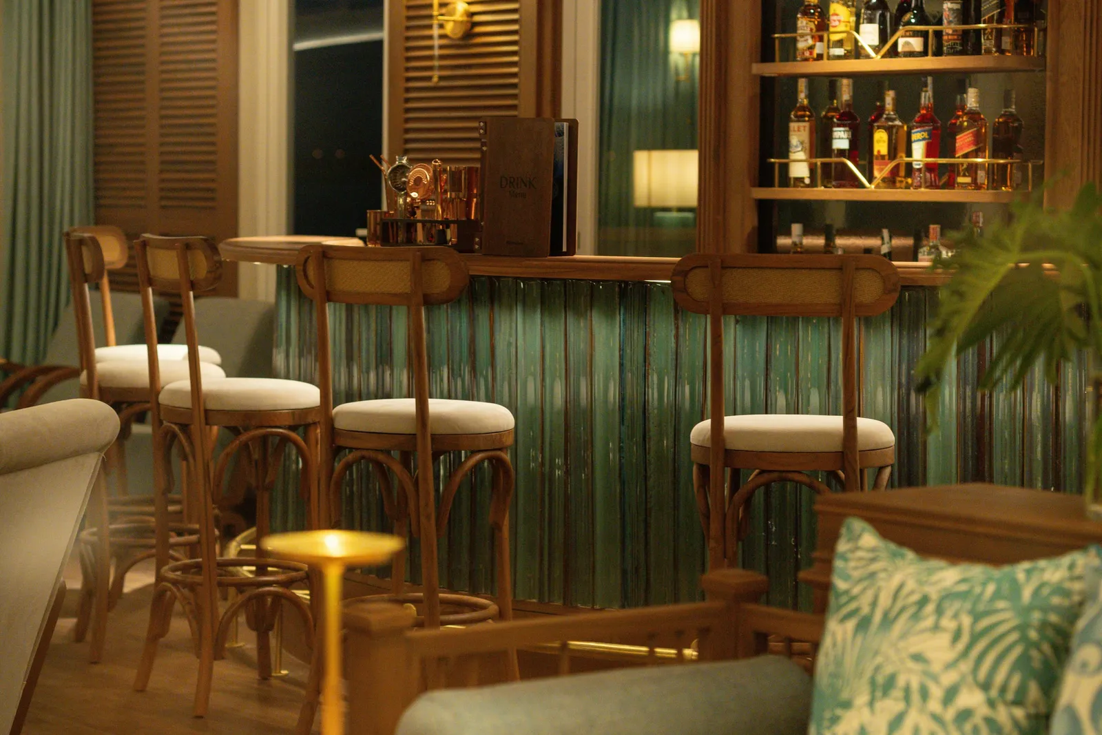 A warmly lit bar interior with wooden bar stools in front of a green glass-fronted counter, a bottle shelf behind, a menu folder reading 'DRINK' on the counter, and a patterned cushion and plant in the foreground.