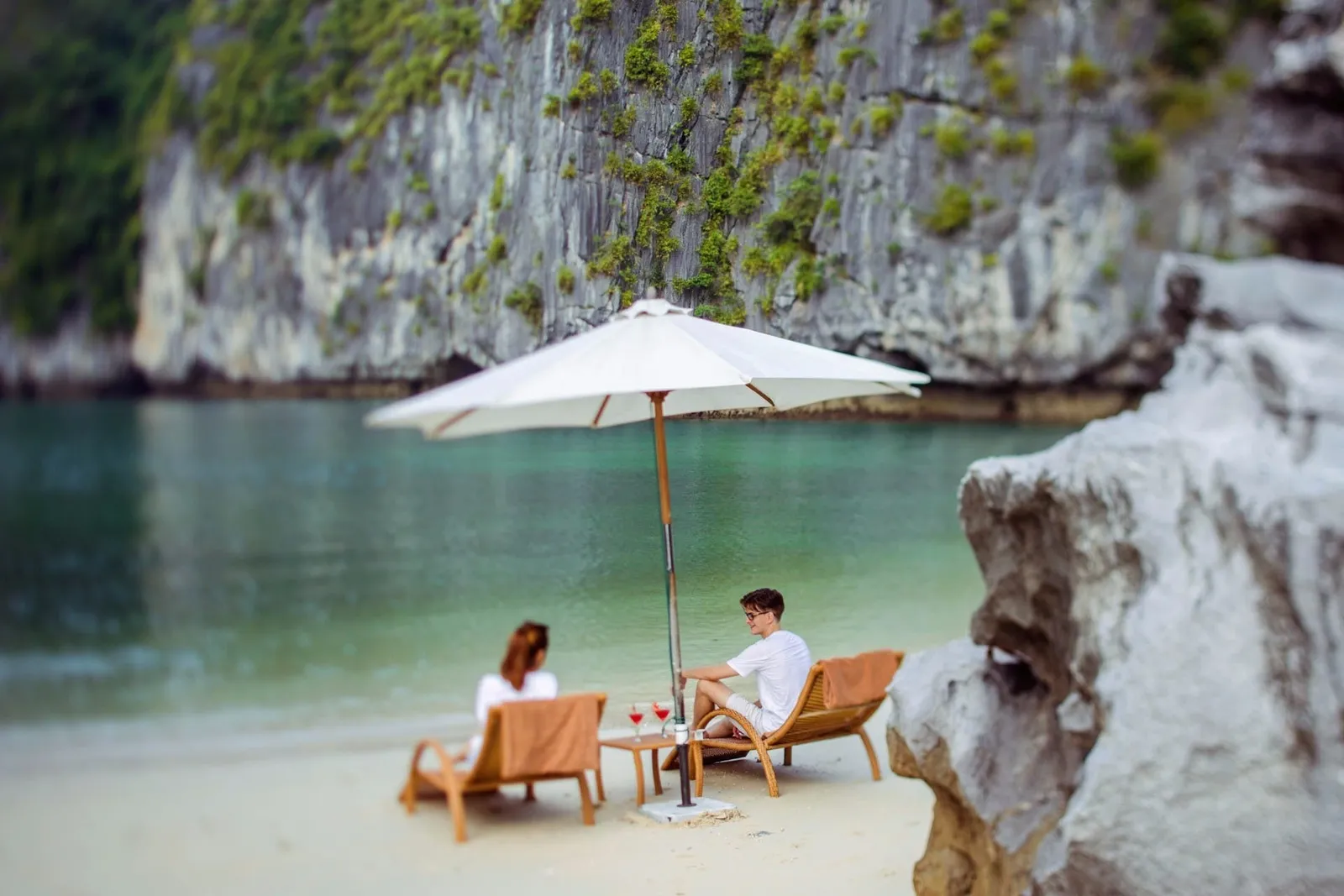 Two people in white sit on wooden loungers with towels under a white parasol on a sandy beach beside green water, with limestone cliffs behind and a rock in the foreground.