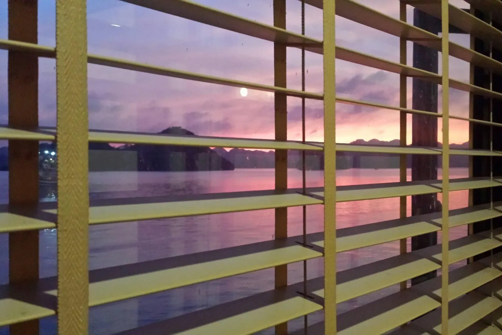 A sunset view of calm water and distant limestone karst hills with a pink and purple sky, seen through a window with wooden blinds.