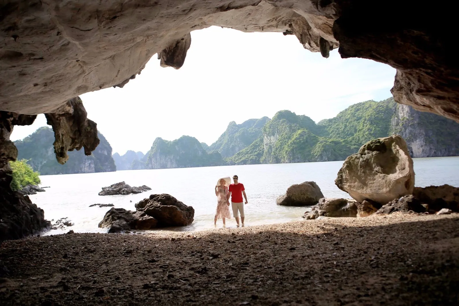 Two people stand hand in hand in shallow water on a small beach, framed by the rocky opening of a cave, with green limestone karst islands across the bay behind them.