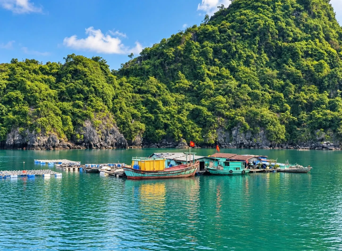 A floating fishing village with colorful wooden boats, fish-farm rafts and small Vietnamese flags sits on turquoise water in front of a steep, forested limestone hill under a blue sky.