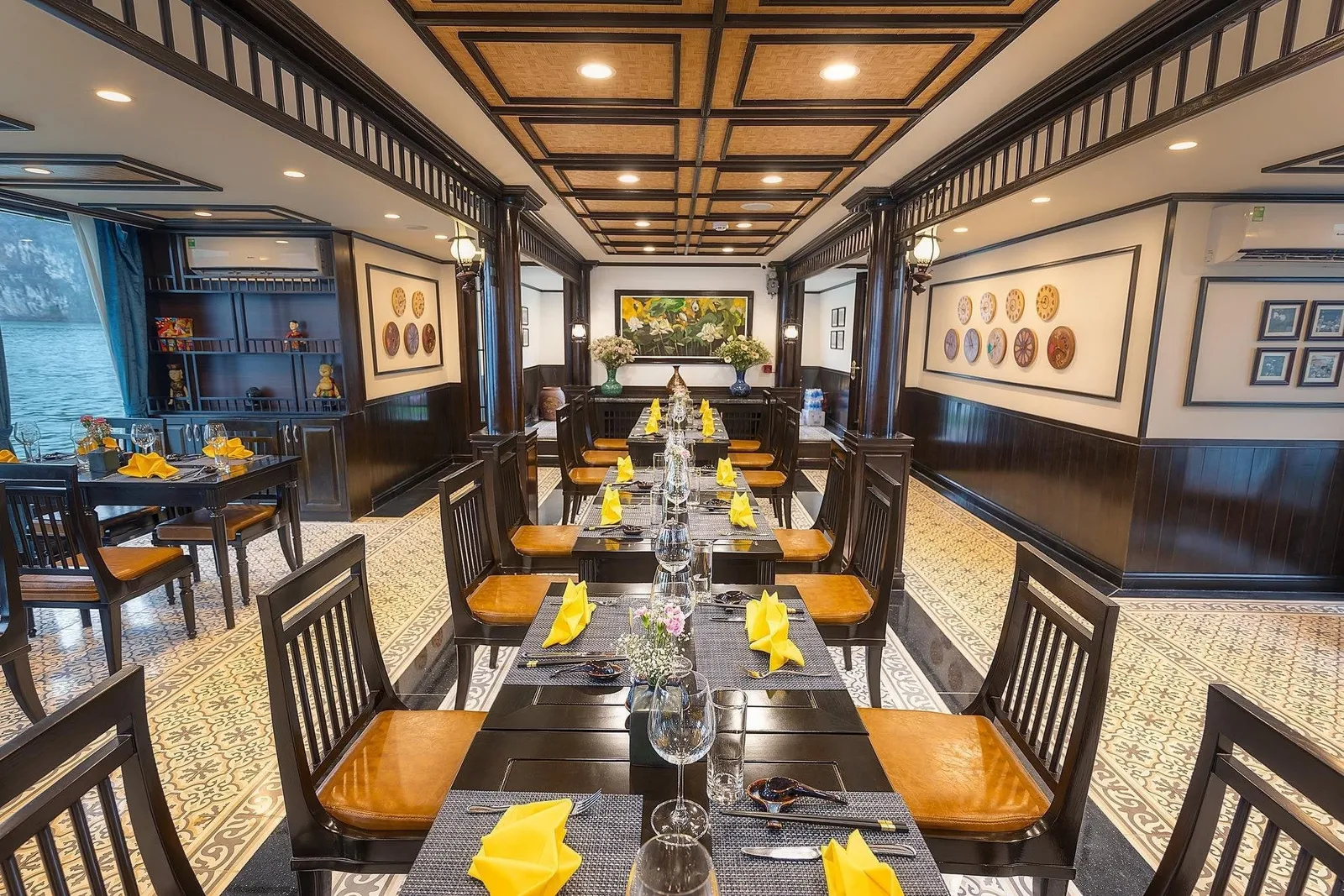 An interior dining room with dark wood tables set with yellow napkins, tan cushioned chairs, patterned floor tiles and decorative wall plates, with a window at left showing water and a rocky karst cliff.