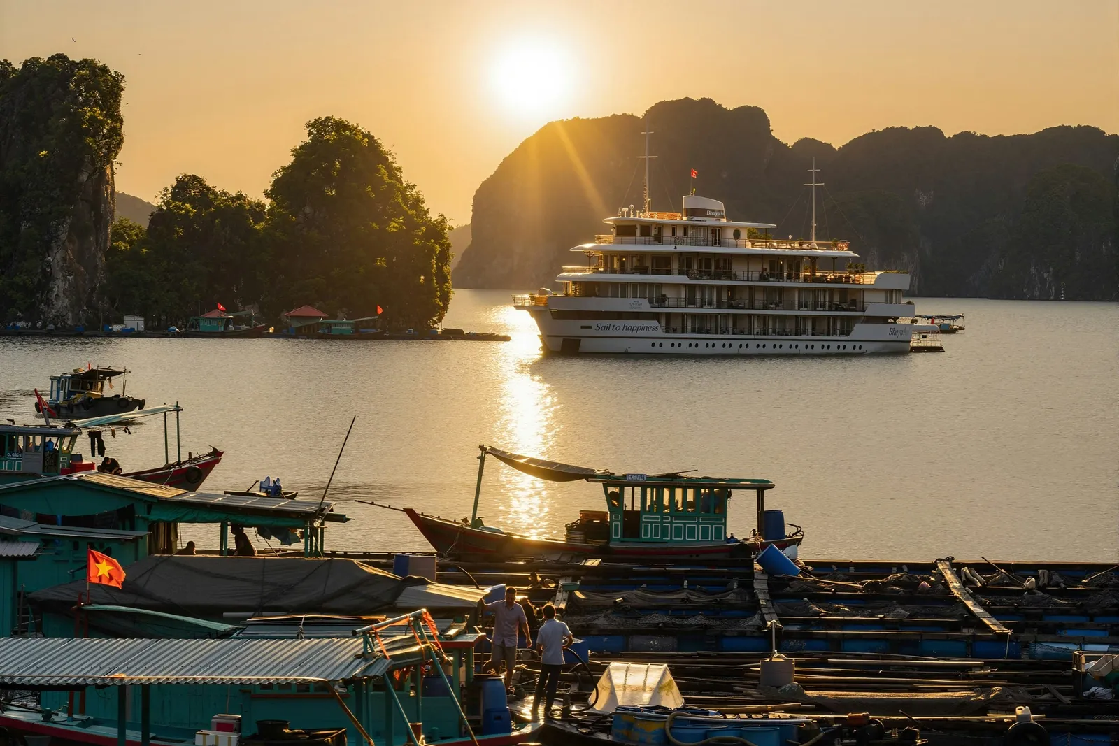 A multi-deck white cruise ship with 'Sail to happiness' on its hull sits on calm water at sunset among limestone karsts, seen from a floating fishing village with small boats, a Vietnamese flag and two people standing on a platform.