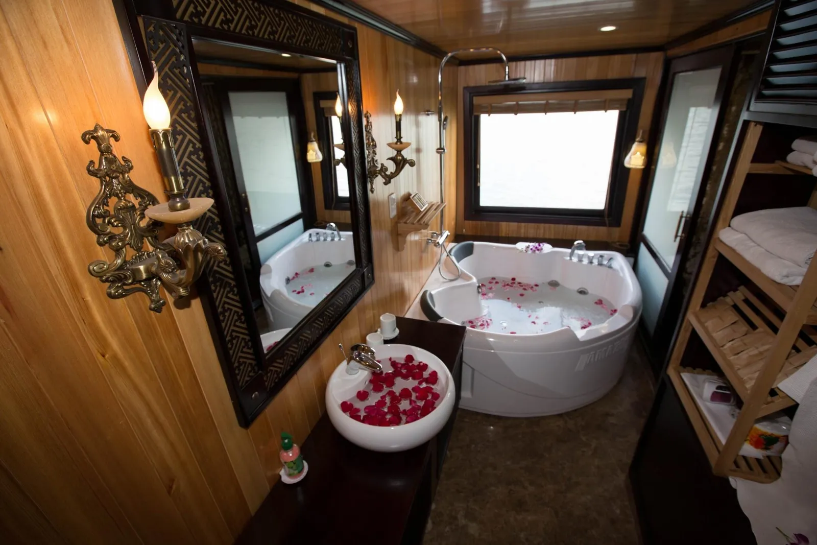 A wood-paneled bathroom with a corner jacuzzi tub and a vessel sink, both filled with rose petals, plus a framed mirror, candle-style wall sconces, a shelf of towels, and a window showing pale water or sky; faint embossed lettering is on the tub's side.
