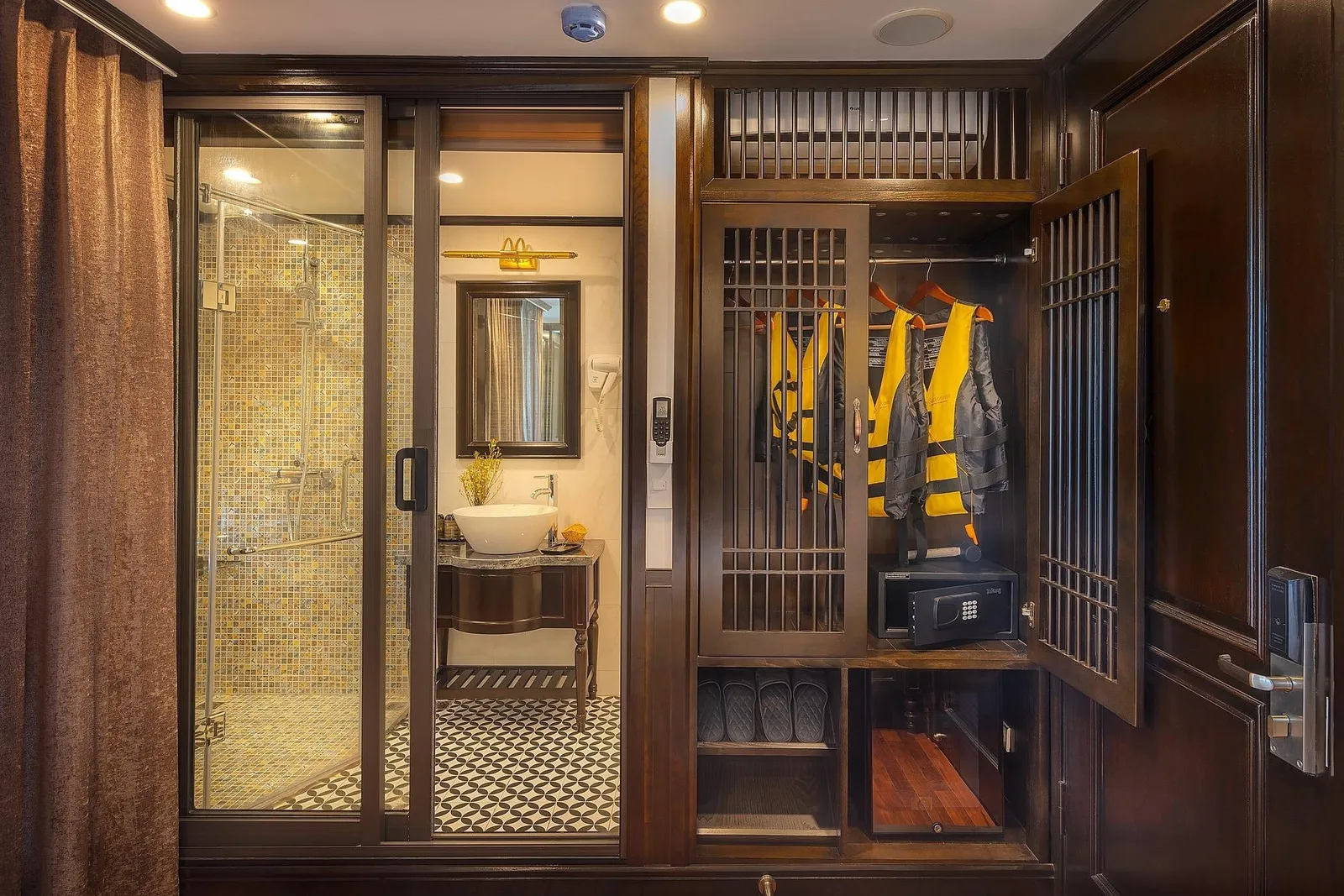 A dark-wood cabin interior with an open wardrobe holding yellow life jackets, a safe and slippers, beside a glass-door shower and a bathroom with a vessel sink and mirror.