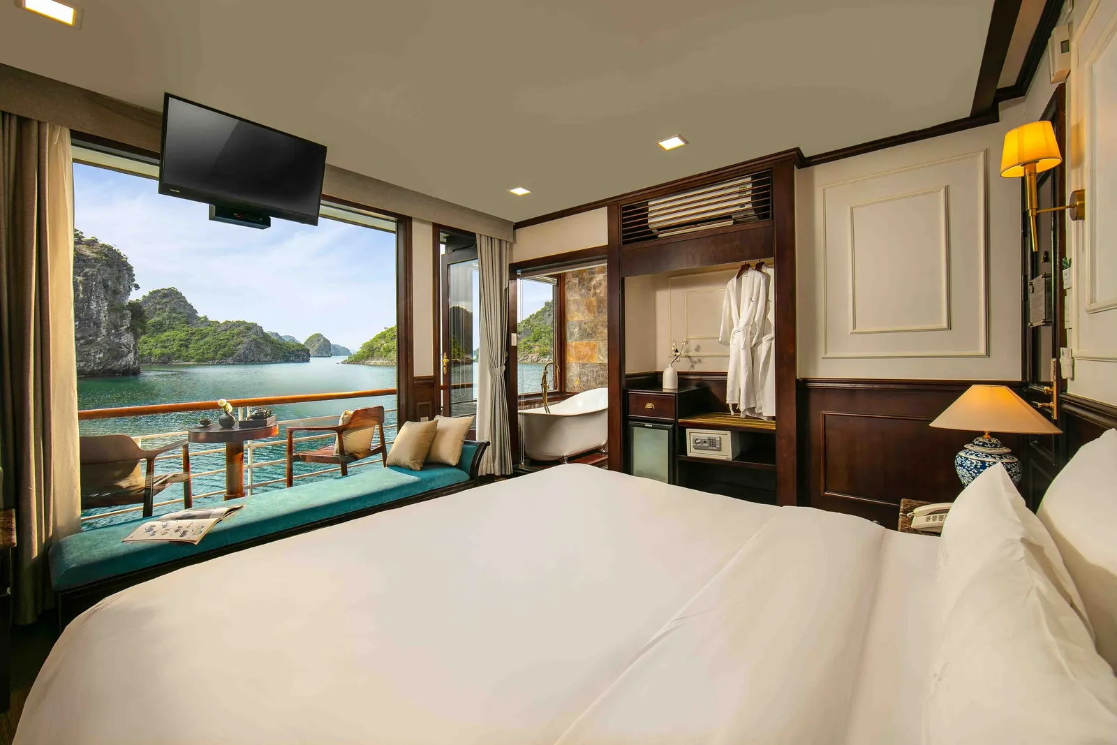 A cabin with a large white bed, wood paneling, a bathtub and hanging robes, and a window and balcony looking out on limestone karst islands in a calm bay.