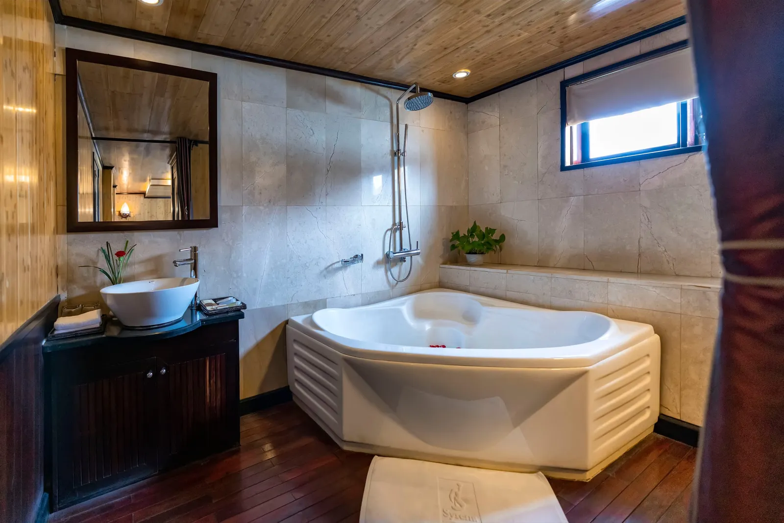 A bathroom with a corner whirlpool tub, a vessel sink on a dark wooden vanity, marble-look tile walls, a wood ceiling and floor, and a bath mat with an embossed logo in the foreground.