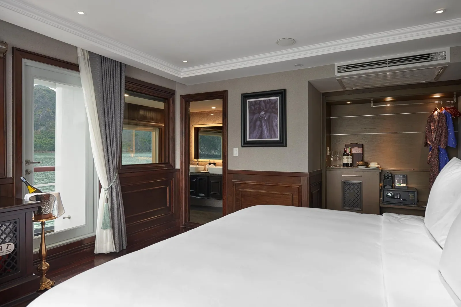A cruise cabin with a large white bed, dark wood paneling, an ensuite bathroom, a wardrobe with robes and a safe, and a balcony door showing green karst hills across the water.