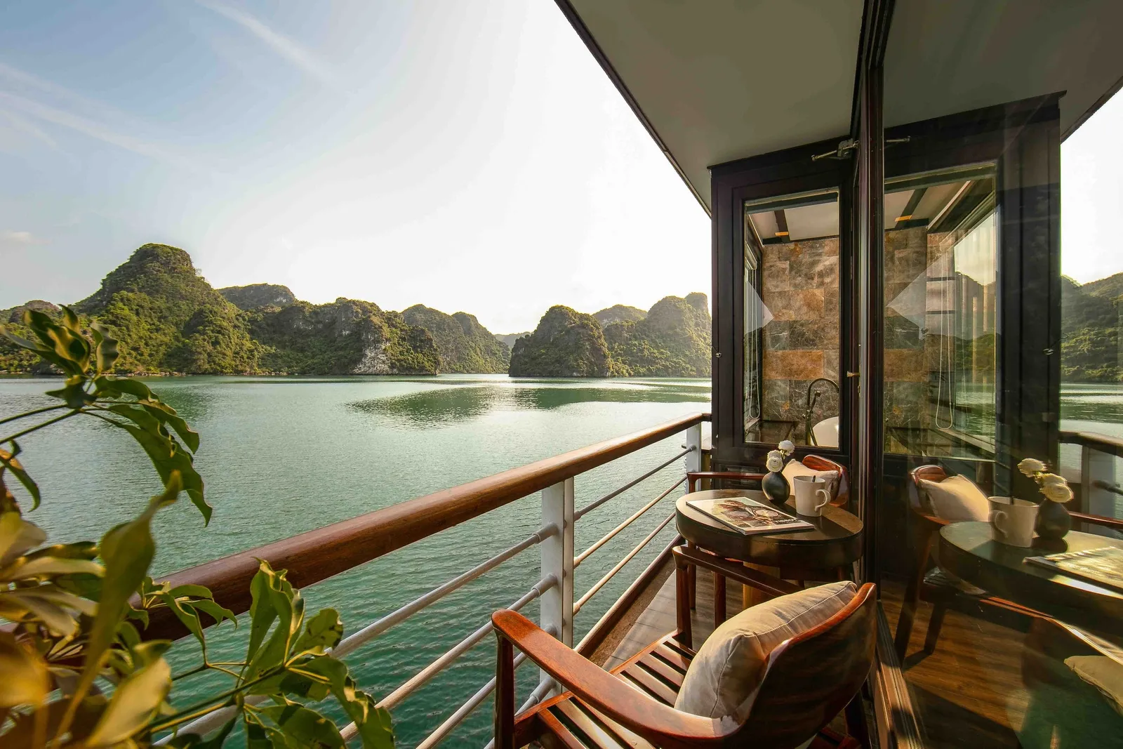 A private wooden balcony on a boat with chairs, a small table with cups and a magazine, a railing, and a glass-walled bathroom, overlooking calm green water and forested limestone karst islands.