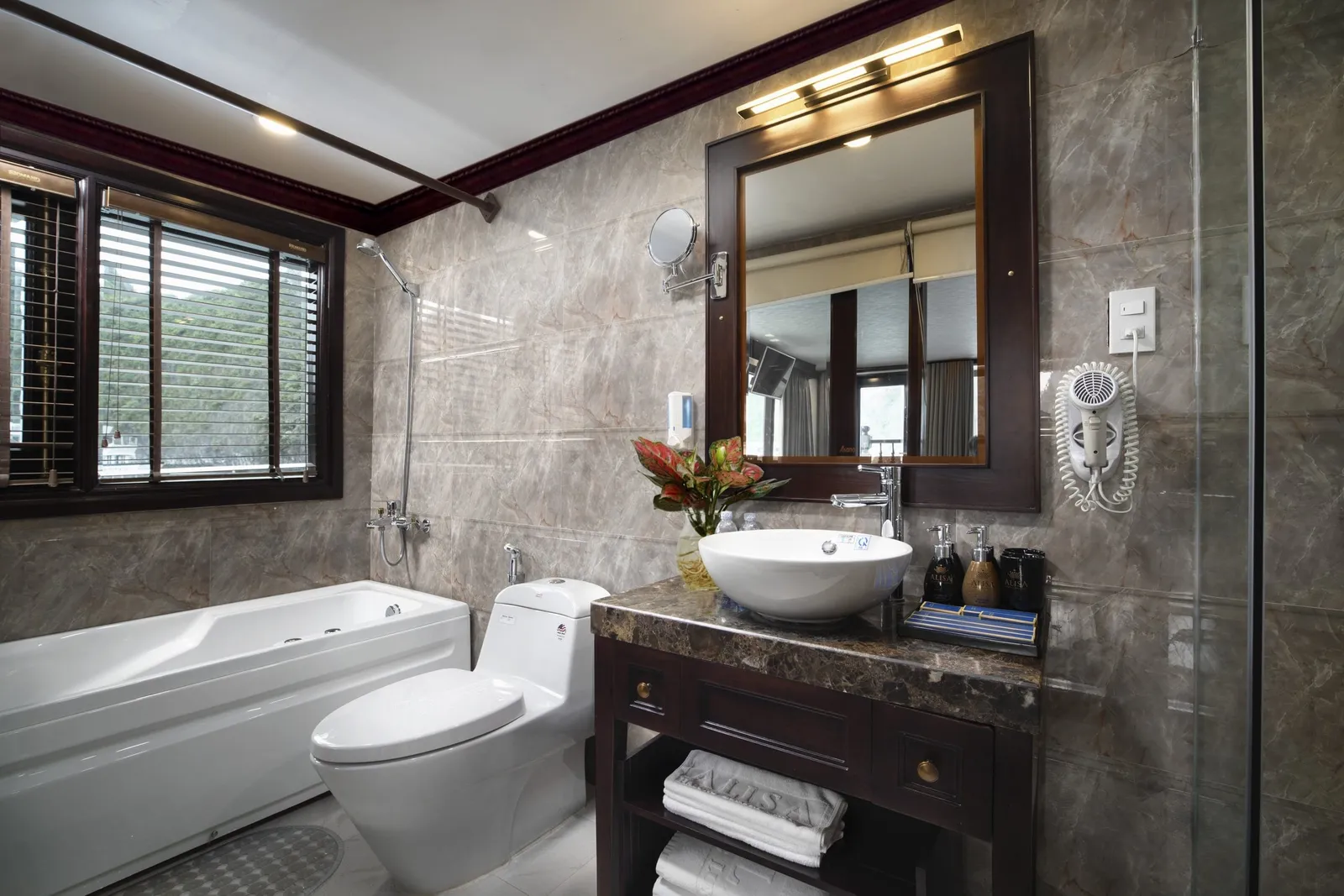 A marble-tiled en-suite bathroom with a bathtub, toilet, vessel sink on a dark wood vanity, framed mirror and a window showing forested limestone hills, with lettering embroidered on the folded towels.