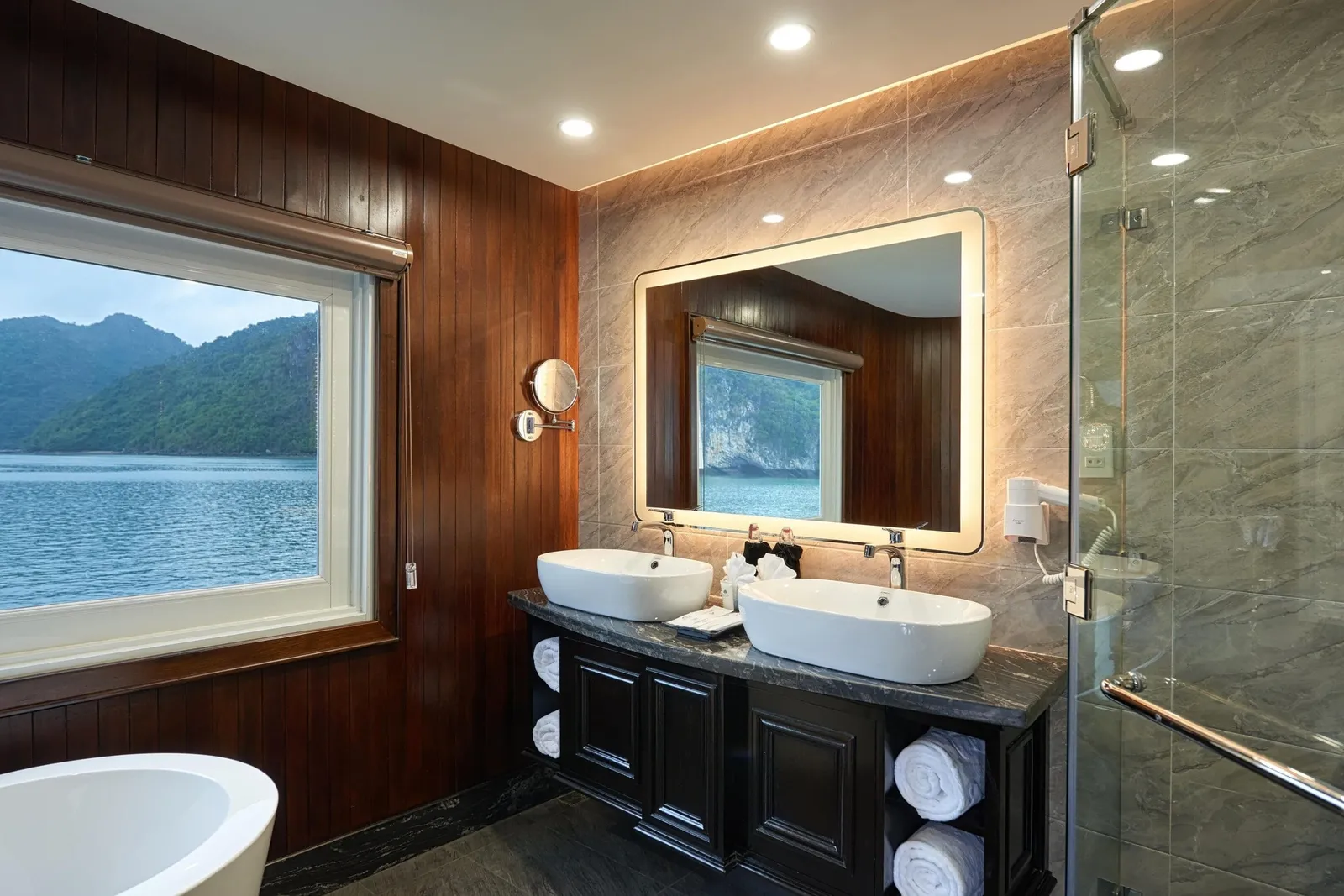 A wood-paneled cruise bathroom with a double vessel-sink vanity, lit mirror, glass shower and freestanding tub, with a window showing calm water and green karst hills.