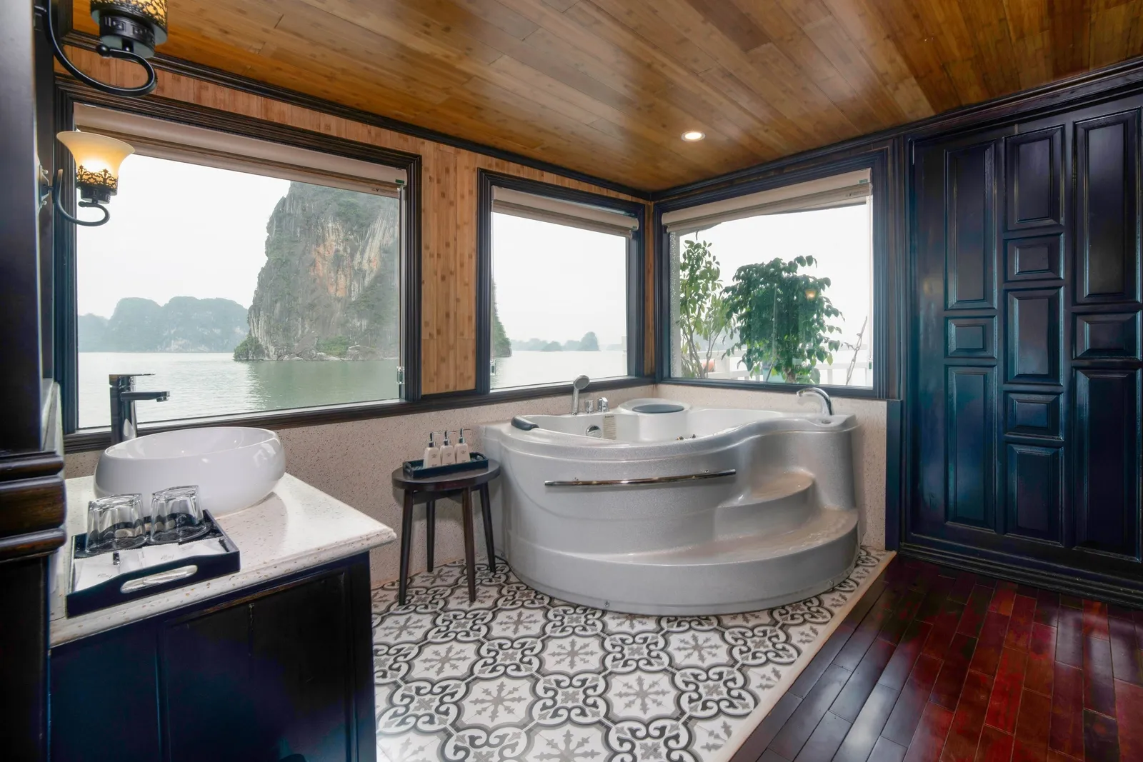 A cabin bathroom with a corner jacuzzi tub, vessel sink, wooden ceiling and dark wood cabinets, with large windows looking out onto calm water and a limestone karst cliff.