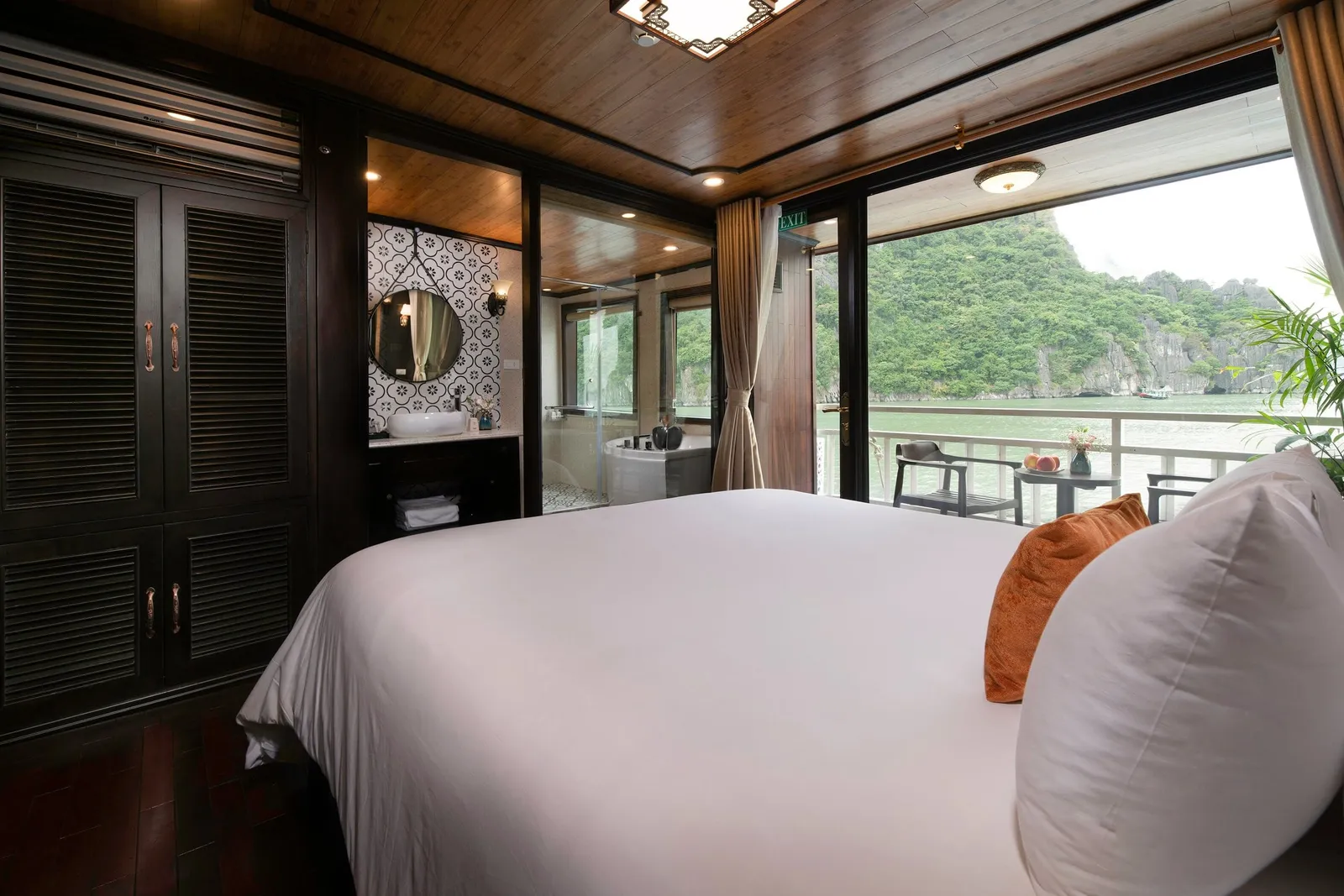 A wood-paneled cruise cabin with a white double bed, orange cushion, open bathroom with tub and round mirror, and glass doors opening to a balcony facing green limestone karst hills over calm water.