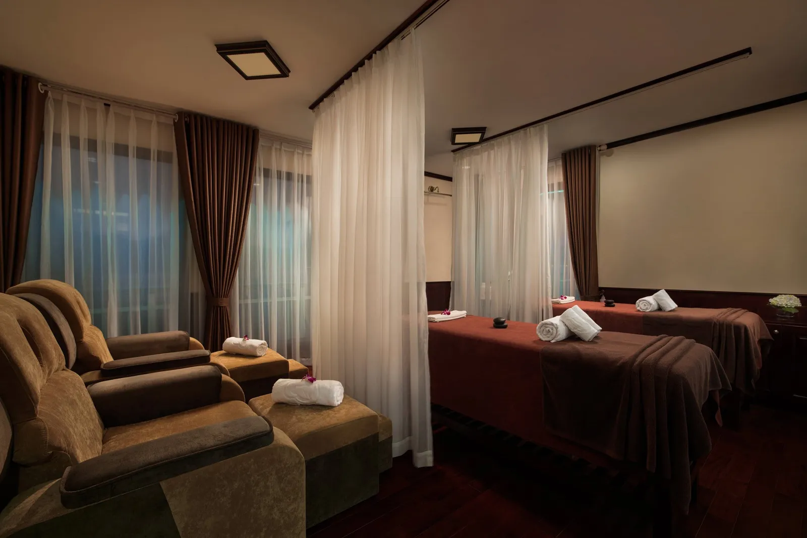A dimly lit spa room with two massage beds with rolled white towels, sheer curtains dividing the space, and two brown upholstered footrest recliners in the foreground.