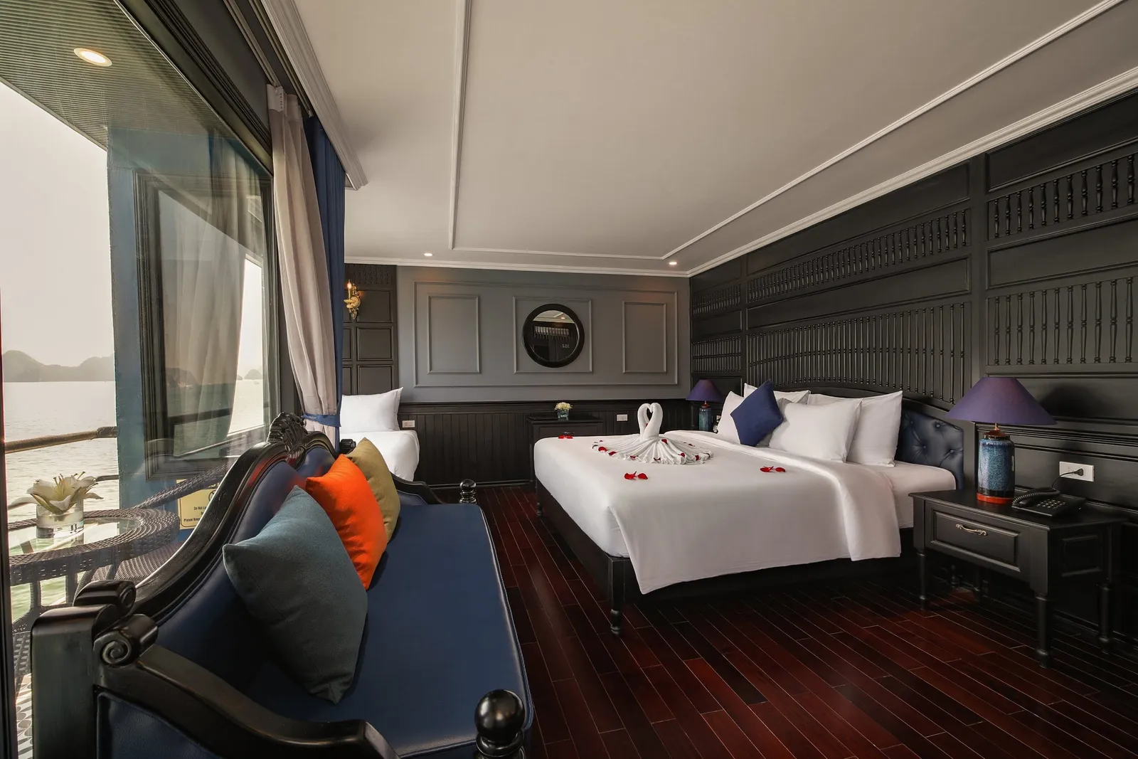 A dark-wood cruise cabin with a double bed decorated with a towel swan and red petals, a blue sofa with colorful cushions, and a large window showing misty water and distant karst islands.