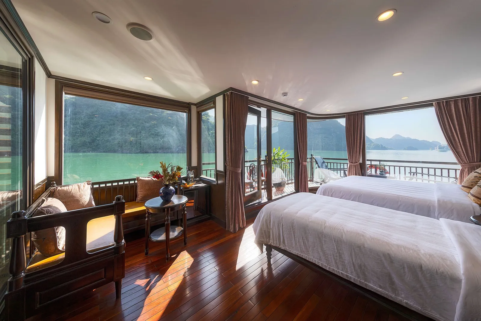 A wood-floored cruise cabin with two twin beds, a carved bench seat and small round table with flowers, and large windows and glass doors opening onto a balcony with green water and forested limestone hills outside.